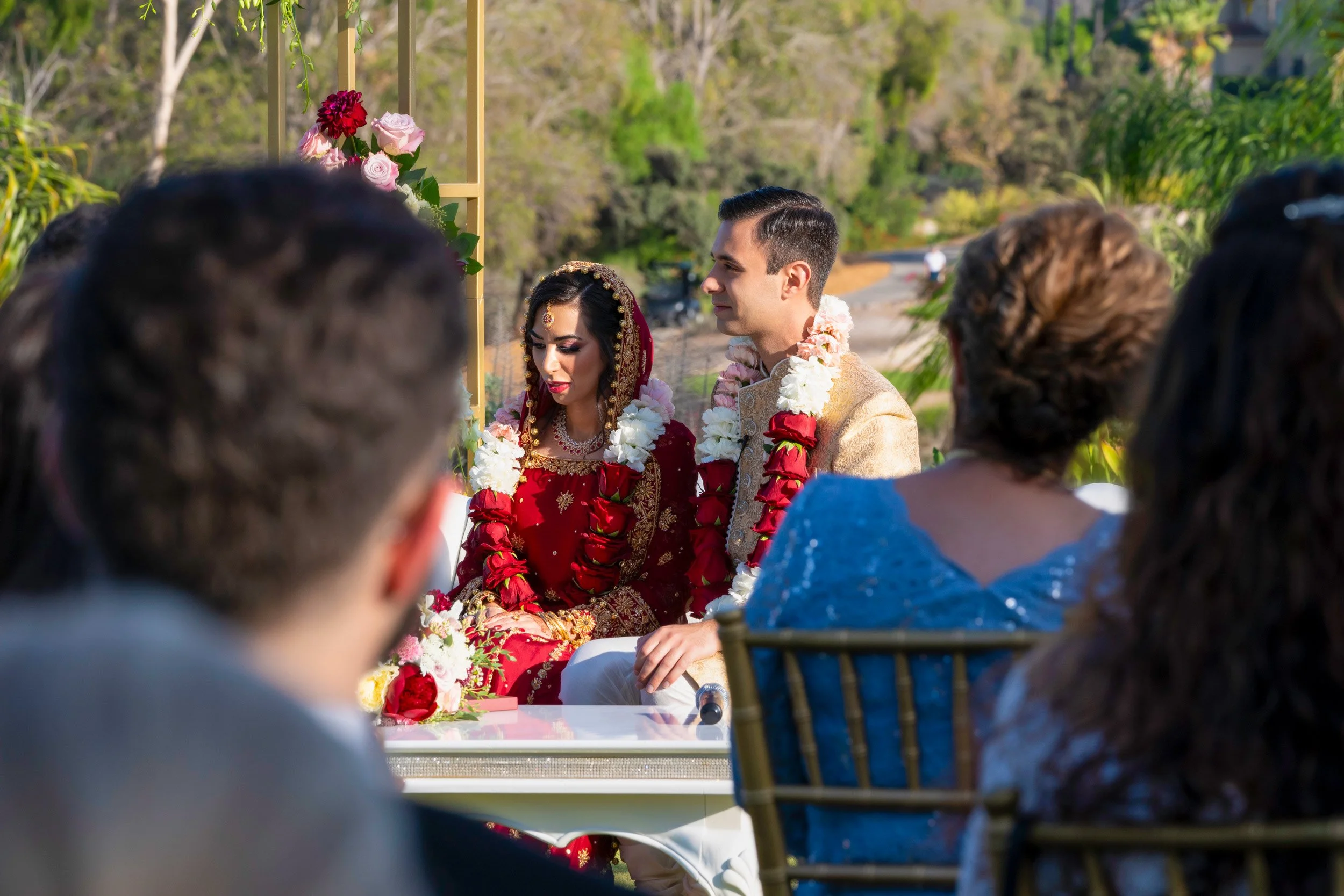An outdoor photograph capturing a bride and groom seated at their wedding altar during a ceremony. The bride wears a traditional deep red lehenga with gold accents and a veil, while the groom wears a gold sherwani. Both are adorned with red and white