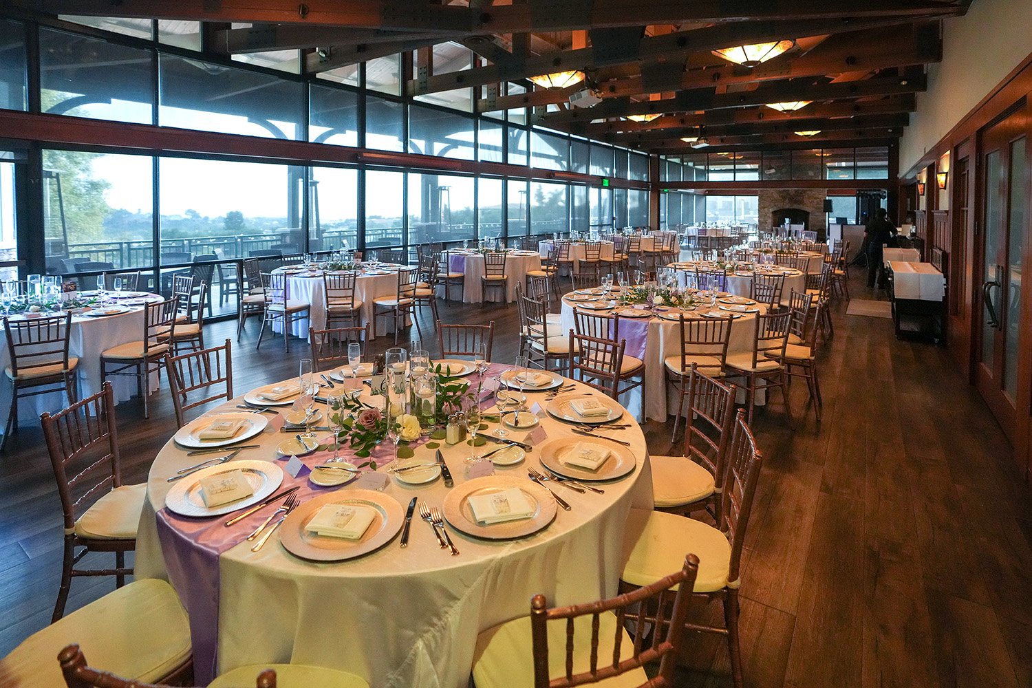 A large, elegant indoor banquet hall or dining room set up for an event, featuring round tables with white and lavender linens, gold charger plates, gold chiavari chairs, exposed wooden beams on the ceiling, and floor-to-ceiling windows with a view o