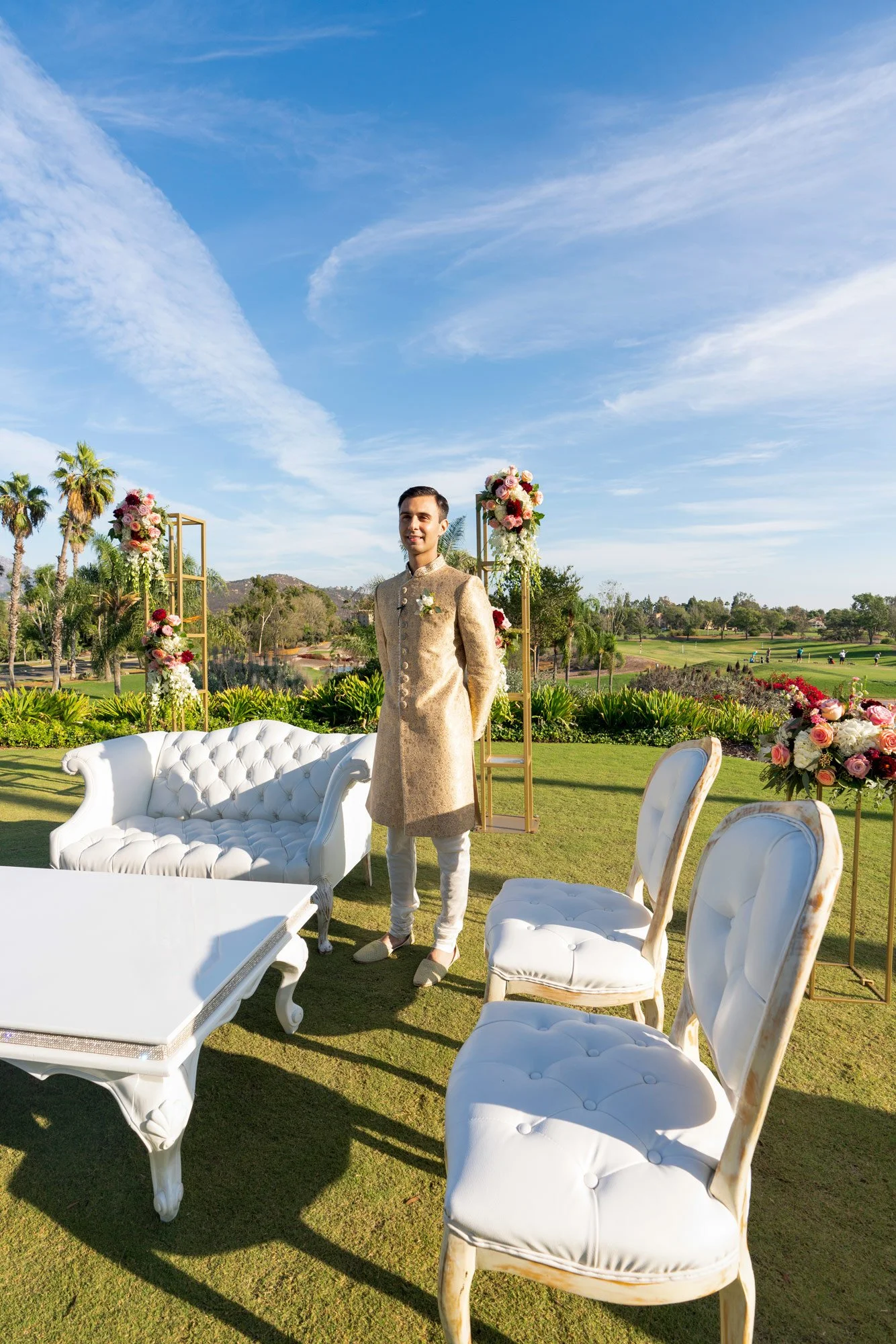 An outdoor photograph of a groom dressed in a traditional gold sherwani, standing in a decorated wedding ceremony area. He stands in front of a white tufted loveseat, between floral arrangements on gold stands, on a sunny day with a blue sky, appeari