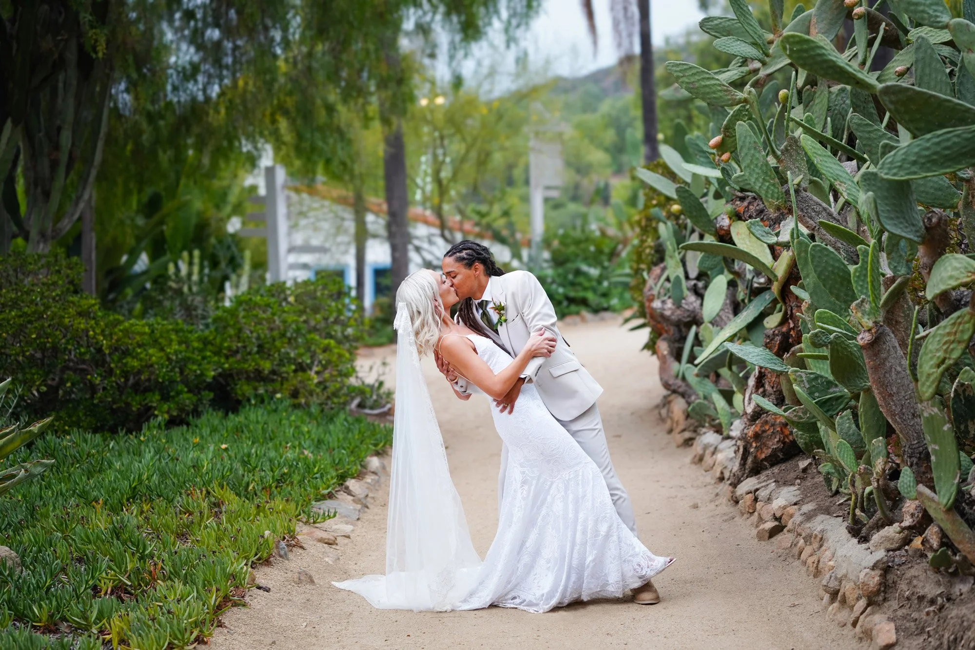 A photo of a newly married couple kissing on a dirt path, surrounded by lush green plants and large prickly pear cacti at Leo Garrillo Ranch Historic Park.