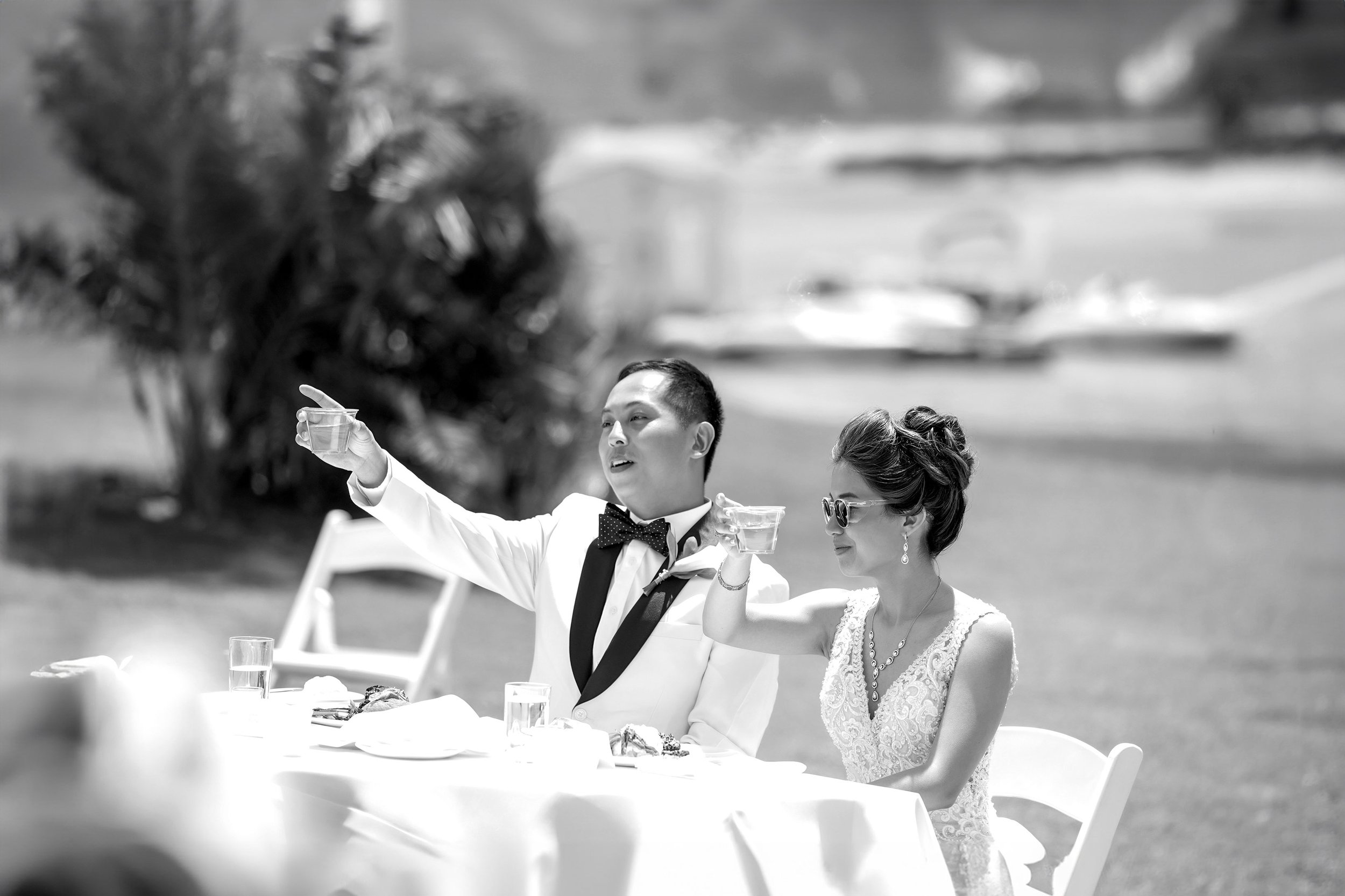 A black and white photograph captures a bride in a lace gown and sunglasses and a groom in a white tuxedo jacket seated at an outdoor table, raising small glasses in a toast or acknowledgment during their wedding reception.