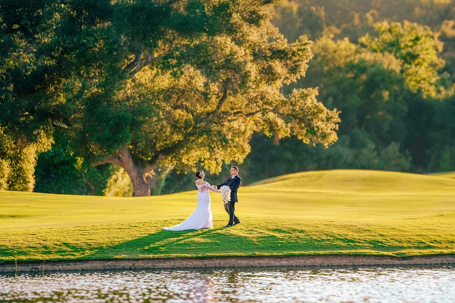 A full-length outdoor photograph of a newly married couple holding hands and looking at each other on a grassy knoll beside a body of water. The bride wears a white wedding dress with a long train and holds a white floral bouquet, while the groom wea