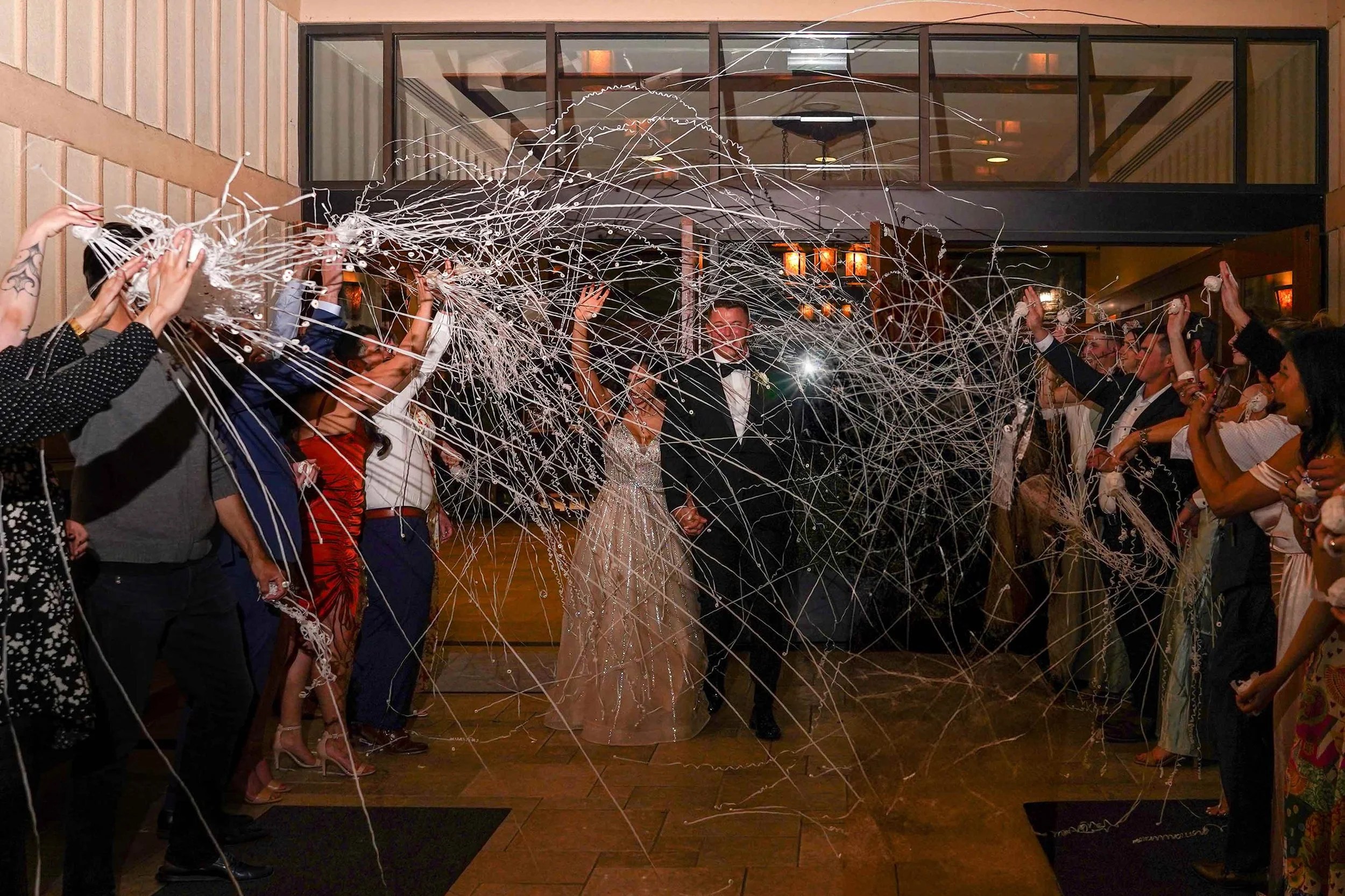An indoor photograph capturing the newly married bride and groom during their reception exit. The couple walks hand-in-hand through a shower of white silly string or streamers being thrown by cheering guests standing on either side of the entranceway