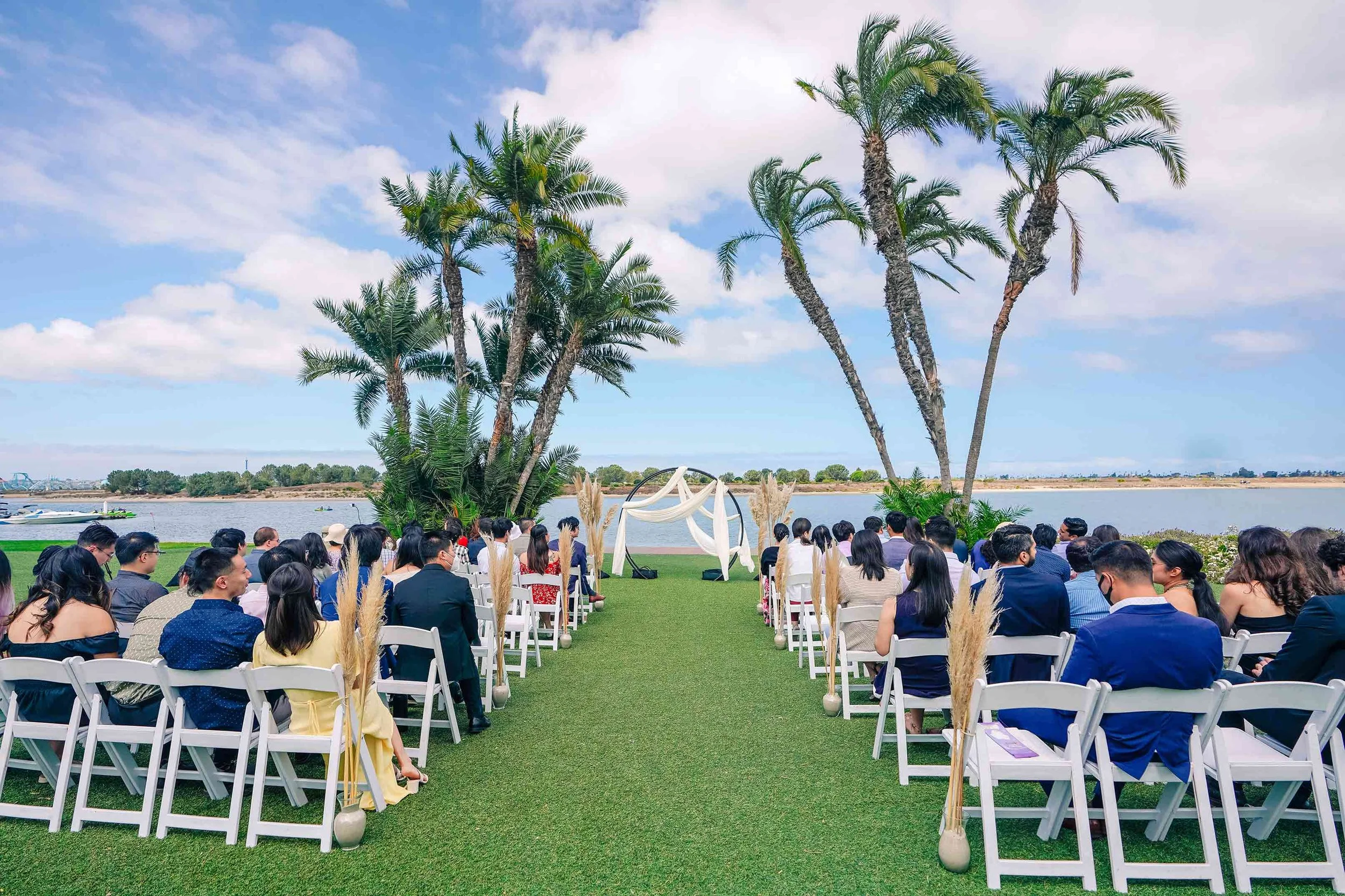 A vibrant, sunny outdoor wedding ceremony set up on the green lawn of Mission Bay, San Diego, featuring seated guests in white chairs, tall palm trees framing an archway, and calm blue water views in the background.