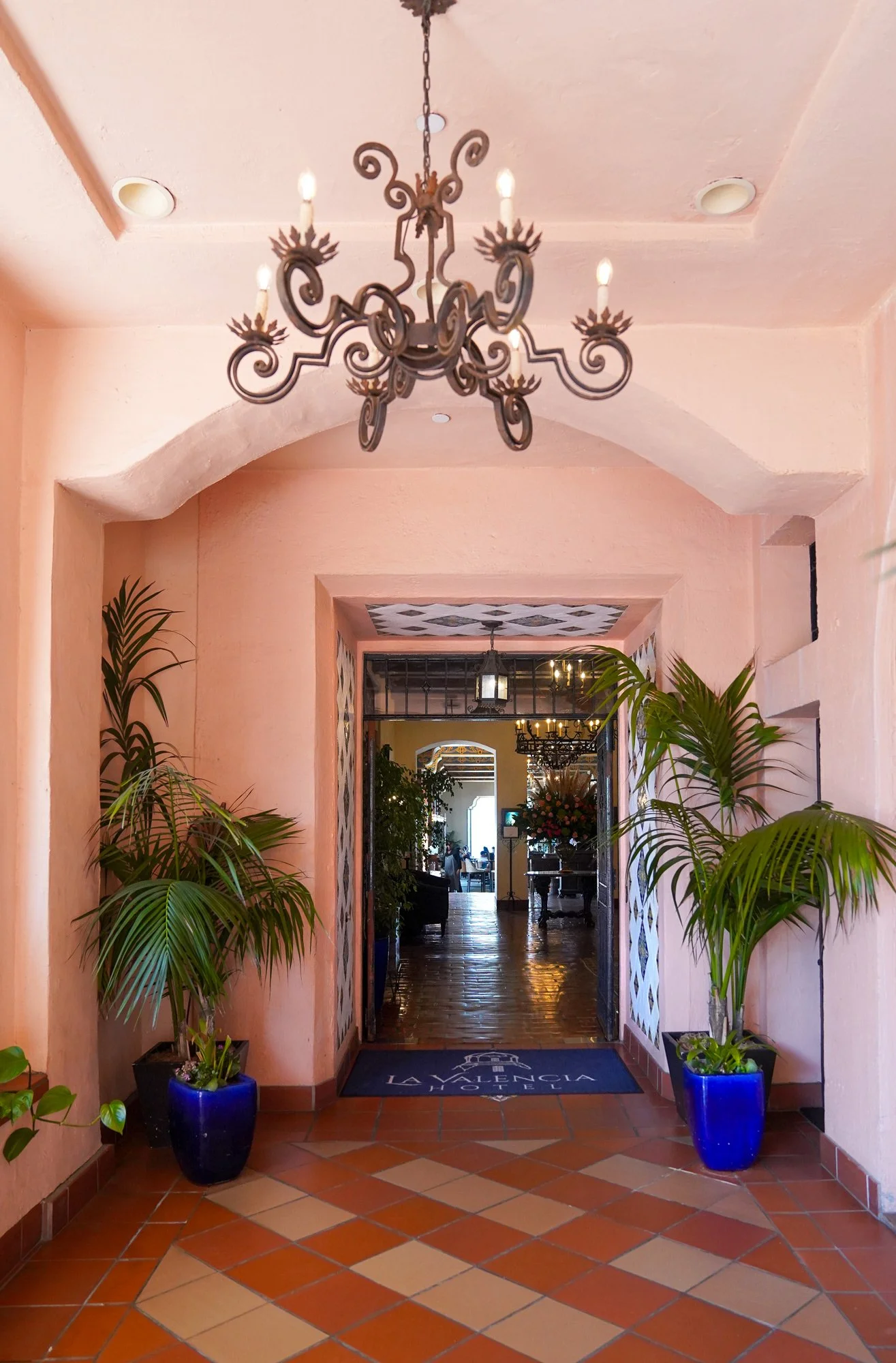 A photograph of an entranceway at the La Valencia Hotel, featuring a terracotta-tiled floor, pink arched walls, and potted palm trees flanking the doorway. An ornate, wrought-iron chandelier hangs from the ceiling, and a doormat with the hotel's name