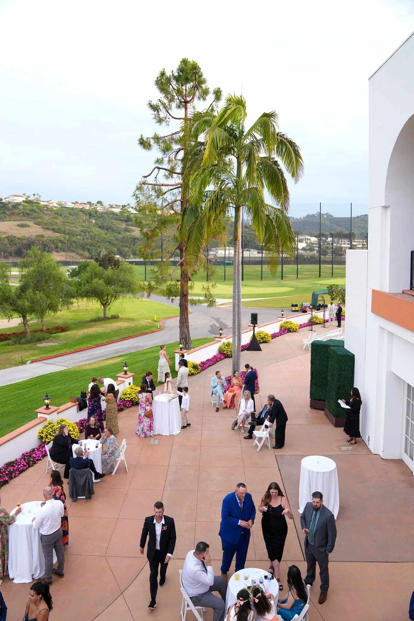 An elevated, daytime view of an outdoor patio event at a resort or country club overlooking a green golf course, with numerous guests mingling at white-clothed tables, two large palm trees, and a white stucco building on the right.
