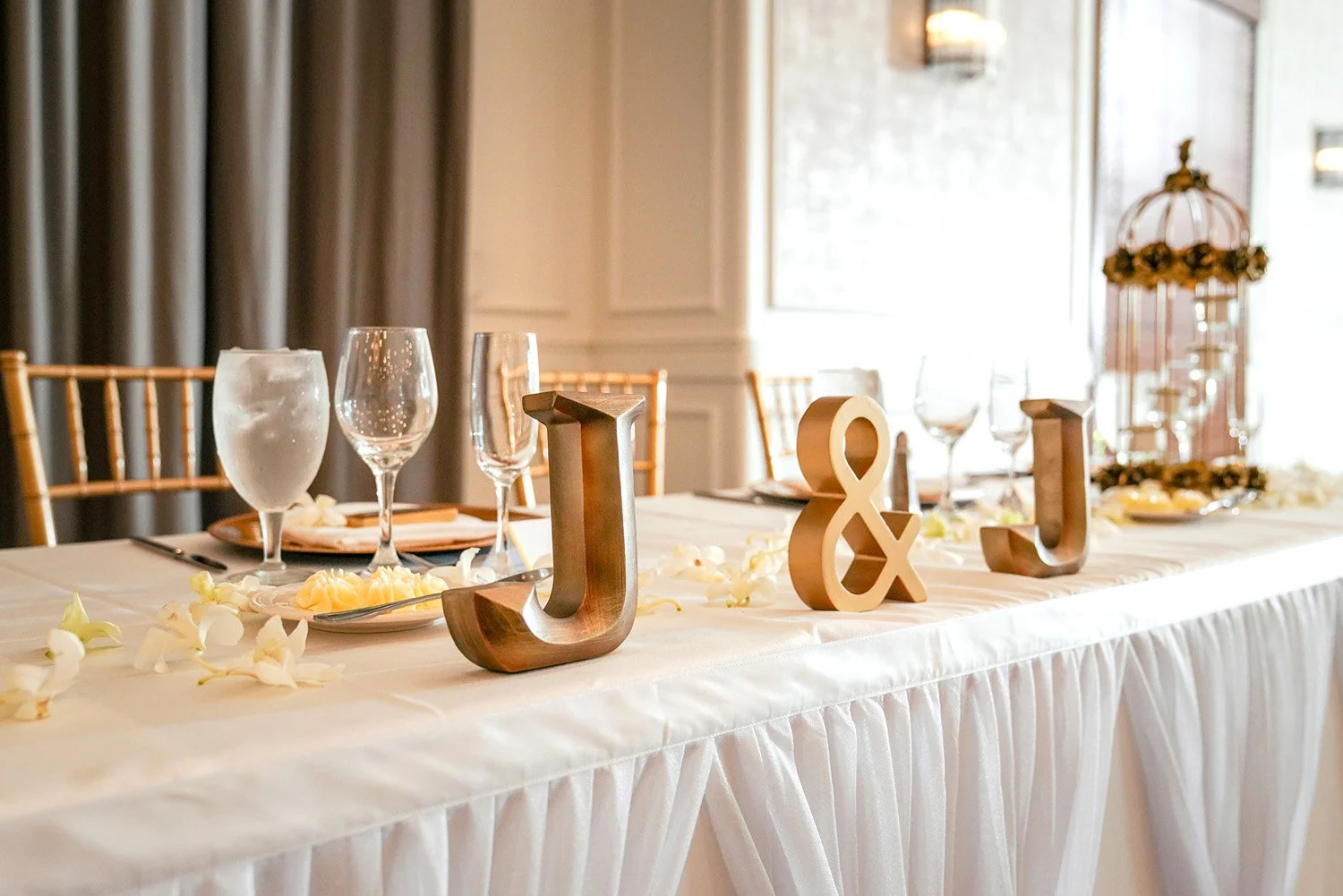 An elegantly set wedding head table, featuring gold "J" and "&" initials as the centerpiece. The table is covered with a white ruffled tablecloth and scattered white flower petals. Place settings include gold charger plates, white plates, and various
