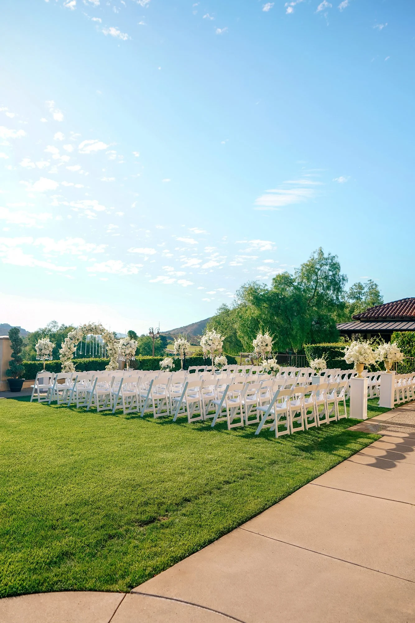 A wide outdoor photograph of an empty wedding ceremony setup on a sunny day. Rows of white folding chairs are arranged on a grassy lawn facing an altar area with a white floral arch and white floral arrangements on pedestals. The background features 