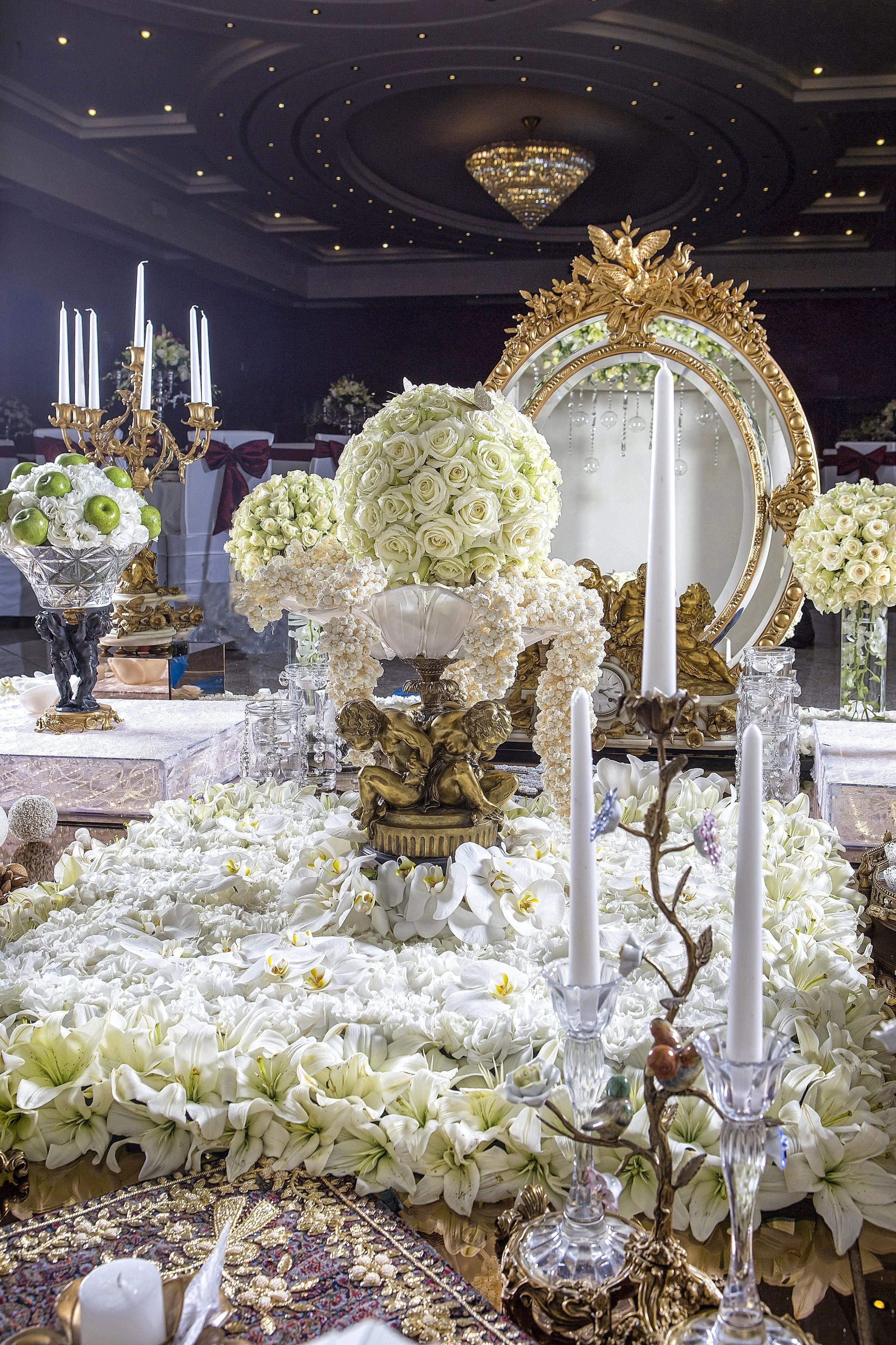 An elaborate, traditional Persian wedding table setting (Sofreh Aghd) featuring a large ornate gold mirror, candelabras, and extensive white floral and gold decorations.
Would you like some suggesti