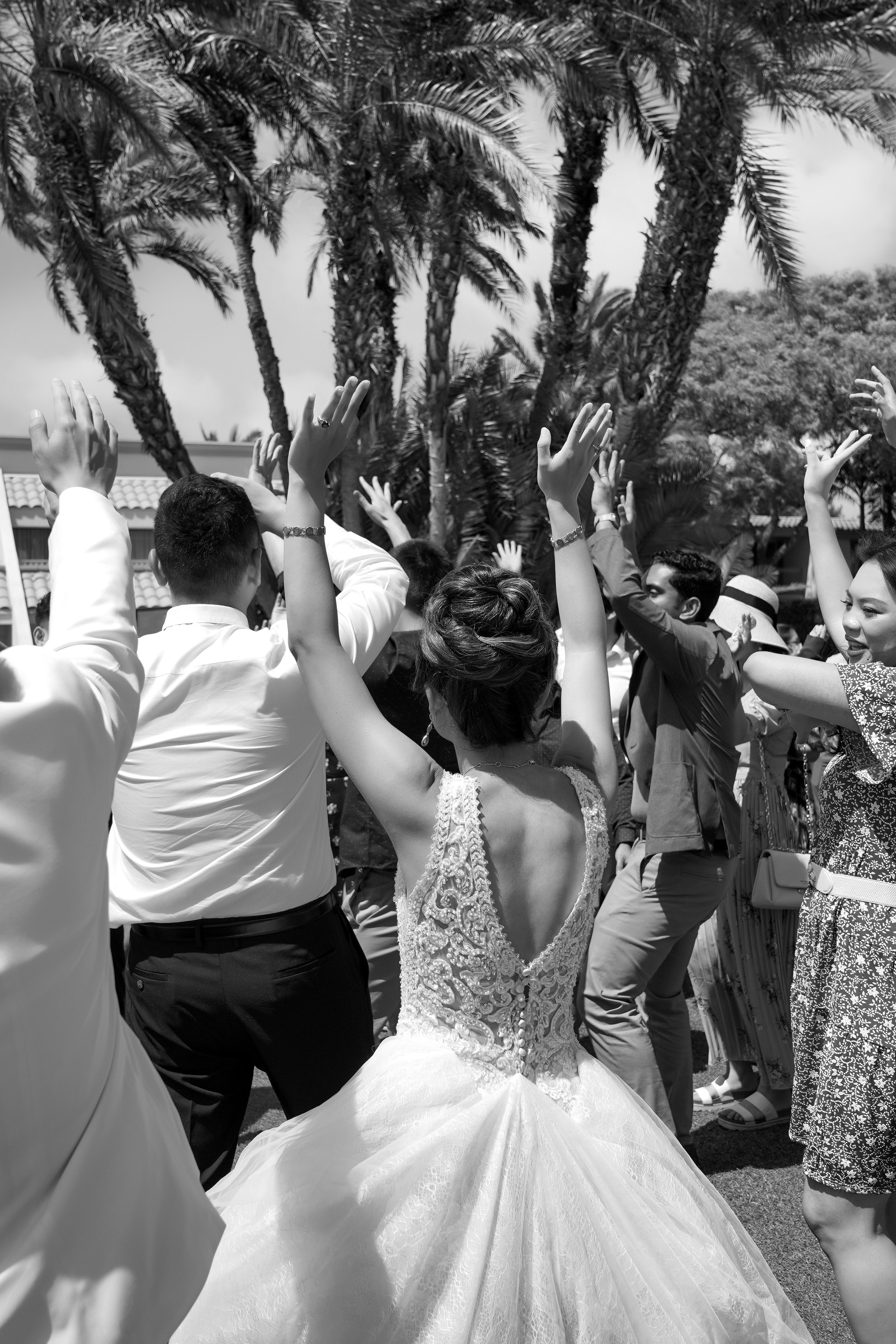 A black and white photo shows a bride with her back to the camera, wearing an ornate lace-top wedding dress and a full tulle skirt. She and a group of guests have their arms raised in celebration outdoors, with palm trees and a clear sky in the backg