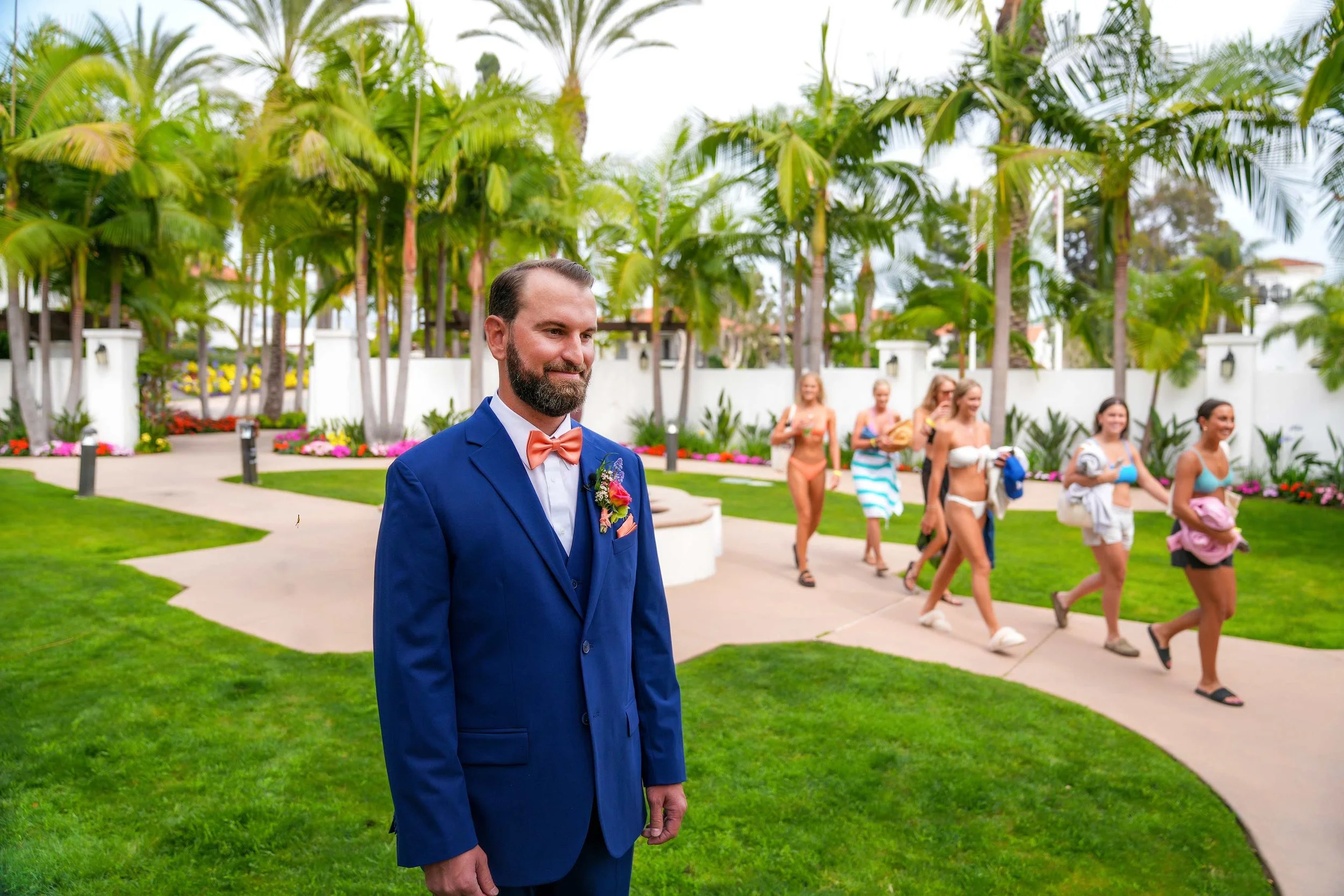 A groom in a blue suit and an orange bow tie stands on a green lawn, looking away from the camera, as a group of young women in swimsuits and casual clothes walk along a paved path at Omni La Costa Resort, surrounded by palm trees in the background.