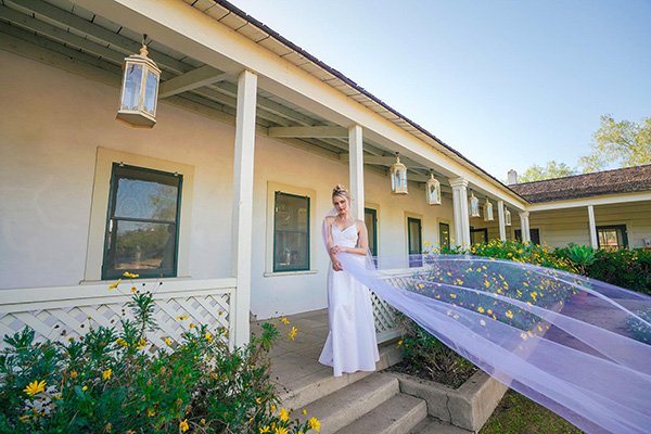 A bride in a white wedding dress stands on a rustic porch, with her long lavender veil catching the wind and flowing across the foreground. 