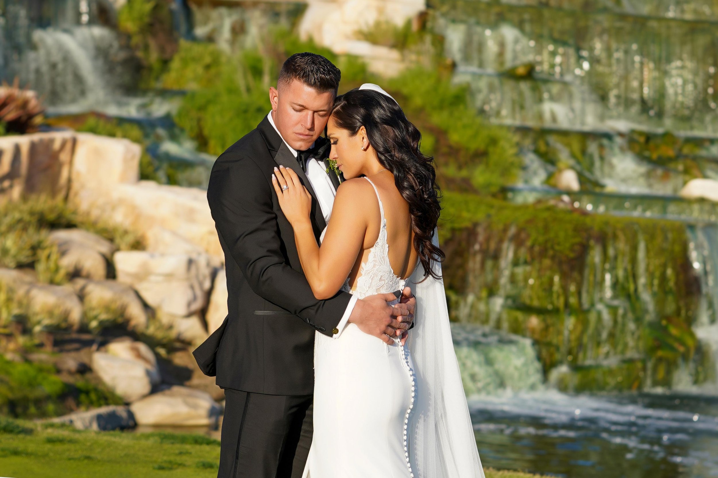 A close-up outdoor photograph of a bride and groom embracing in front of a lush rock waterfall. The groom wears a black tuxedo and bow tie, and the bride wears a white sleeveless lace wedding dress with an open back and a long veil. Both appear to be