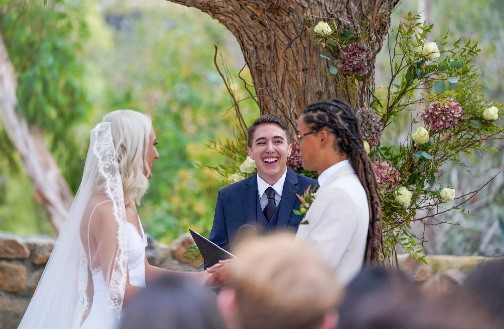 A photo of a wedding ceremony in progress outdoors, with two brides smiling at each other while holding hands in front of an officiant who is laughing. One bride wears a white lace dress and veil, the other wears a white suit, and the officiant is in