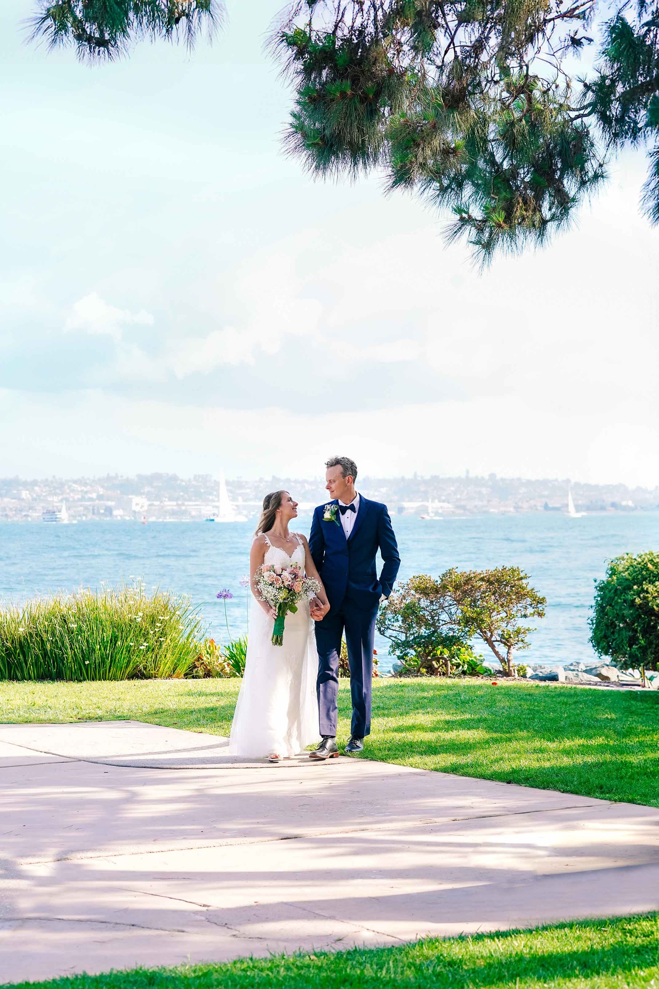 A bride in a white gown and a groom in a navy suit walk along a paved path next to the water, looking at each other in San Diego.


