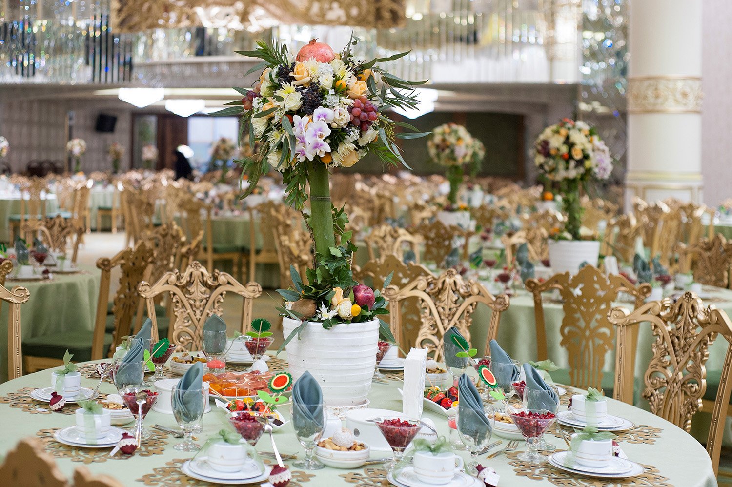 An elegant, formal banquet hall set up for an event, featuring round tables with sage green tablecloths, ornate gold chiavari chairs, and elaborate floral and fruit centerpieces under warm lighting.
