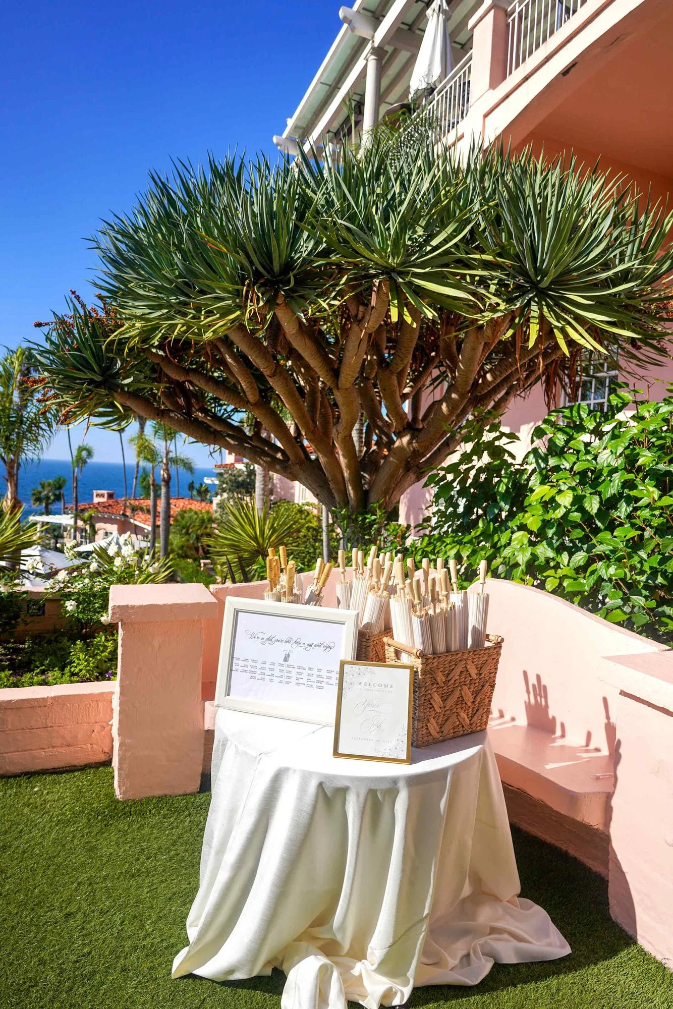 An outdoor photograph of a wedding welcome area on a sunny day, set on a grassy lawn with an ocean view. A white-draped table in the foreground holds a large basket filled with white paper parasols for guests and a framed welcome sign. A large dragon