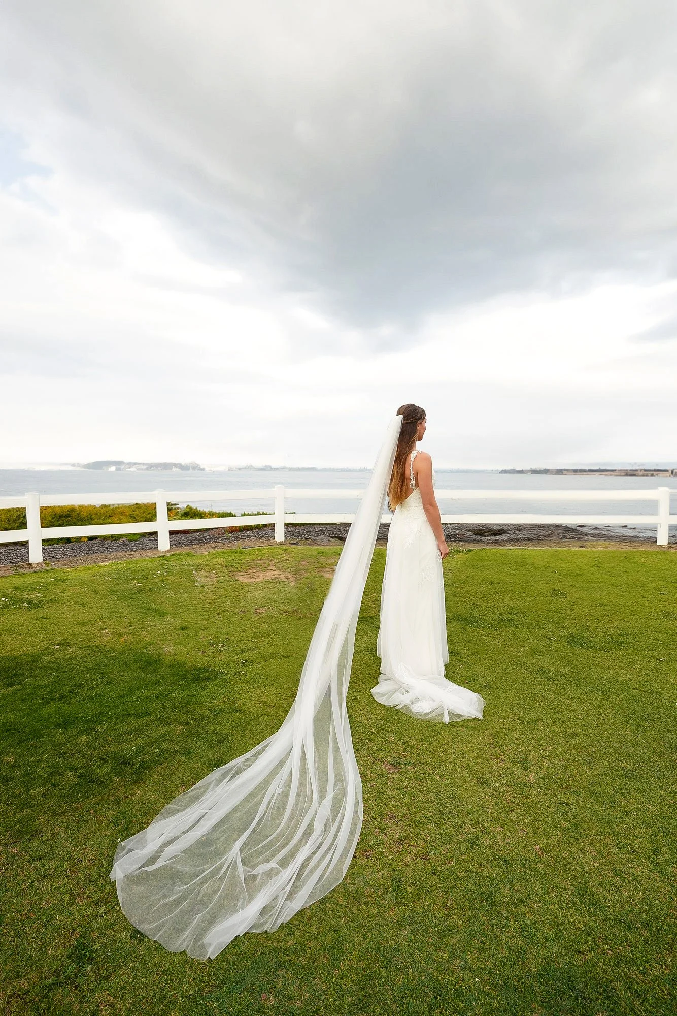 A bride in a white wedding gown and a long, flowing veil stands on a green lawn beside a white fence, looking out over the water and a distant shoreline under a cloudy sky in San Diego.


