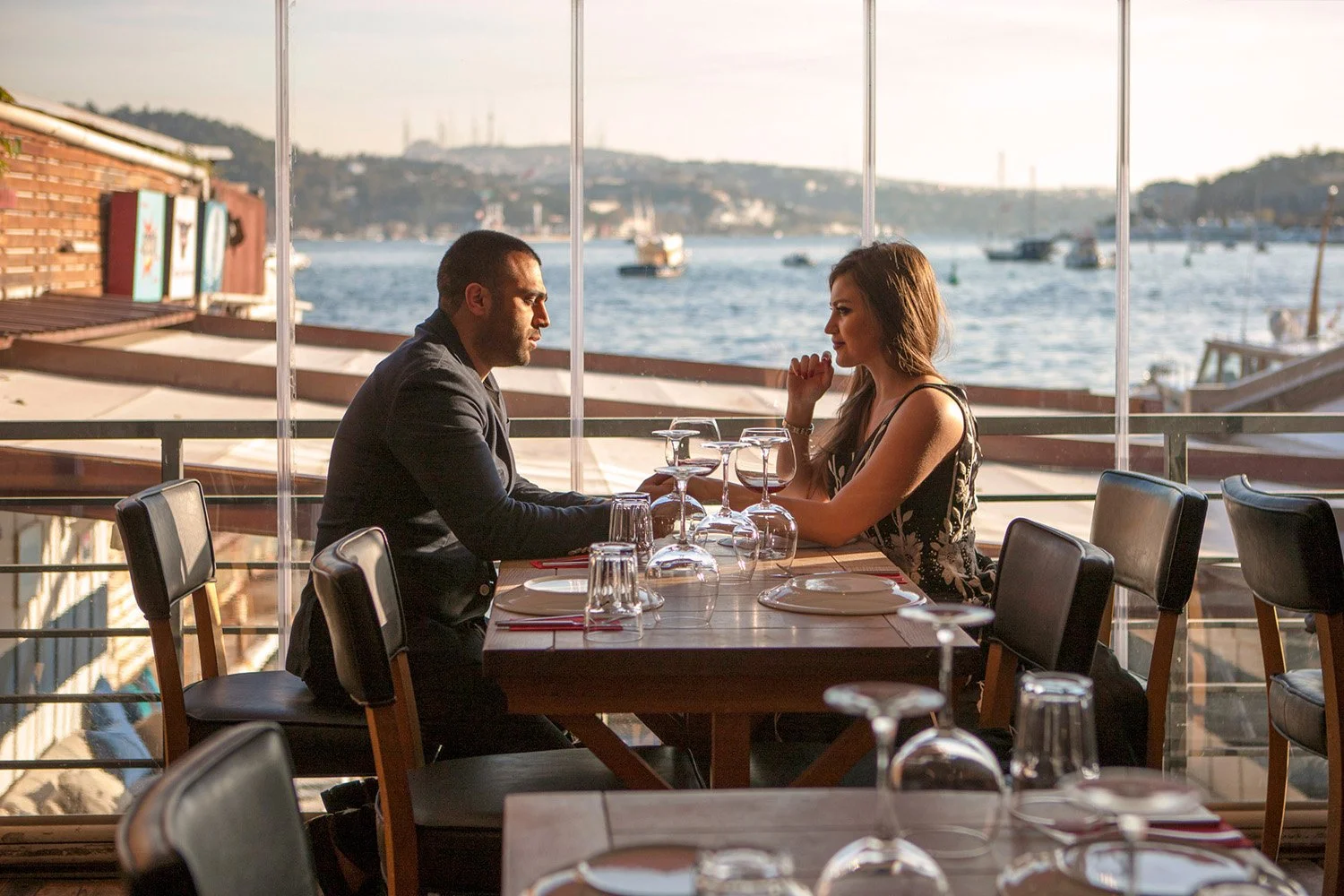 An indoor photograph of a couple having a meal at a restaurant with floor-to-ceiling windows overlooking a body of water and a city skyline of Istanbul. A man in a dark suit sits opposite a woman in a black patterned dress at a wooden table set with 