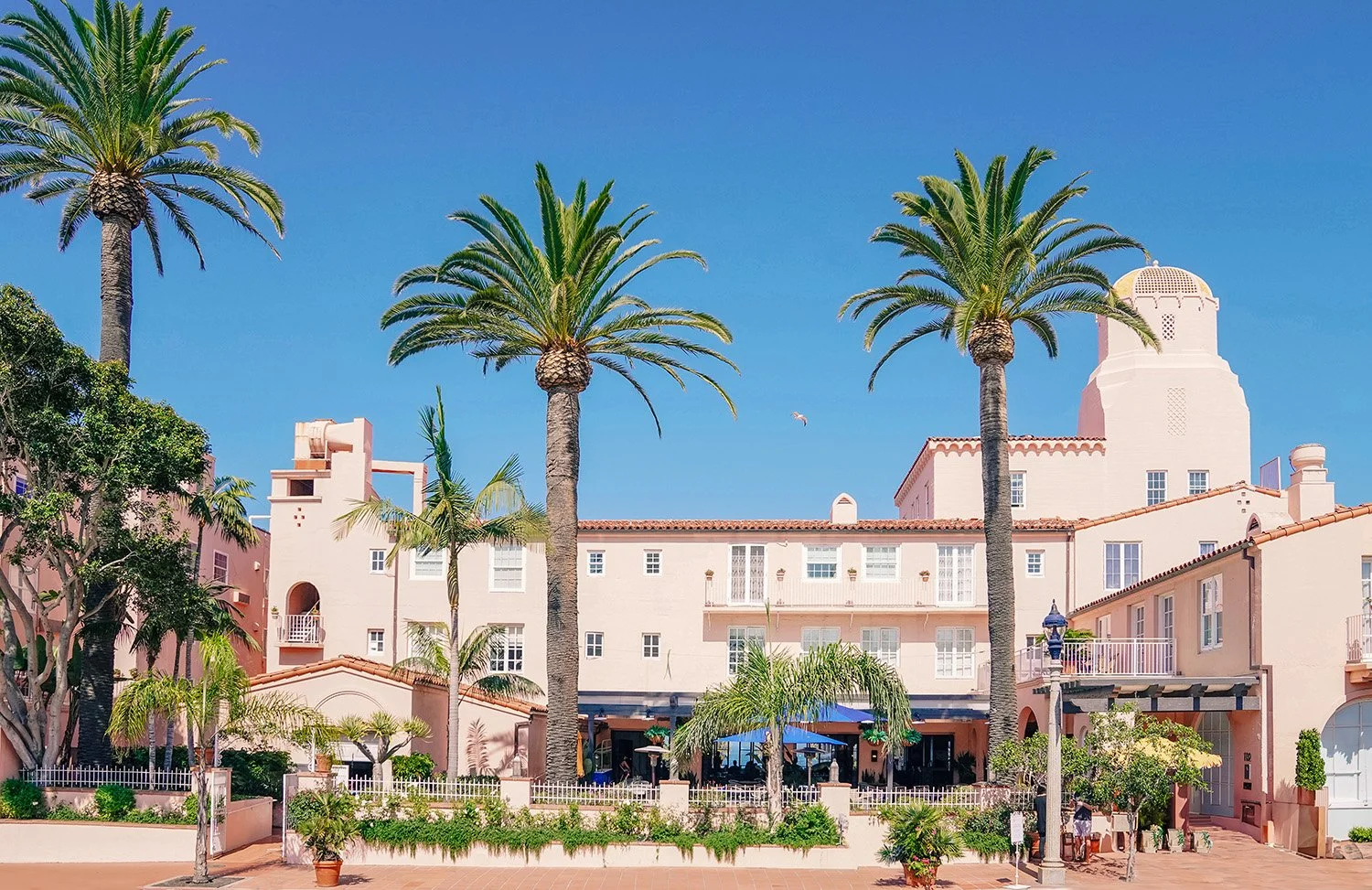 The multi-story, pink-stucco La Valencia Hotel building in La Jolla, California, with a tiled roof, numerous windows, and covered patios framed by large palm trees against a clear blue sky.