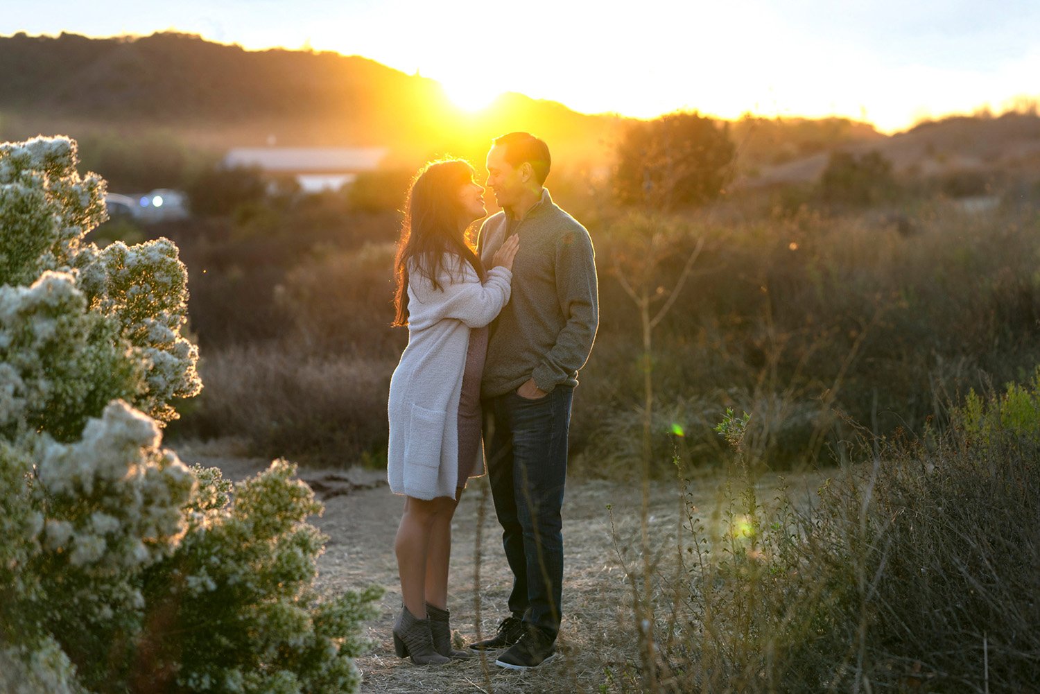A romantic outdoor photograph of a couple embracing and about to kiss during sunset. The bright sun creates a warm lens flare between rolling hills in the background. The couple stands on a dirt path, surrounded by dry golden grasses and green bushes