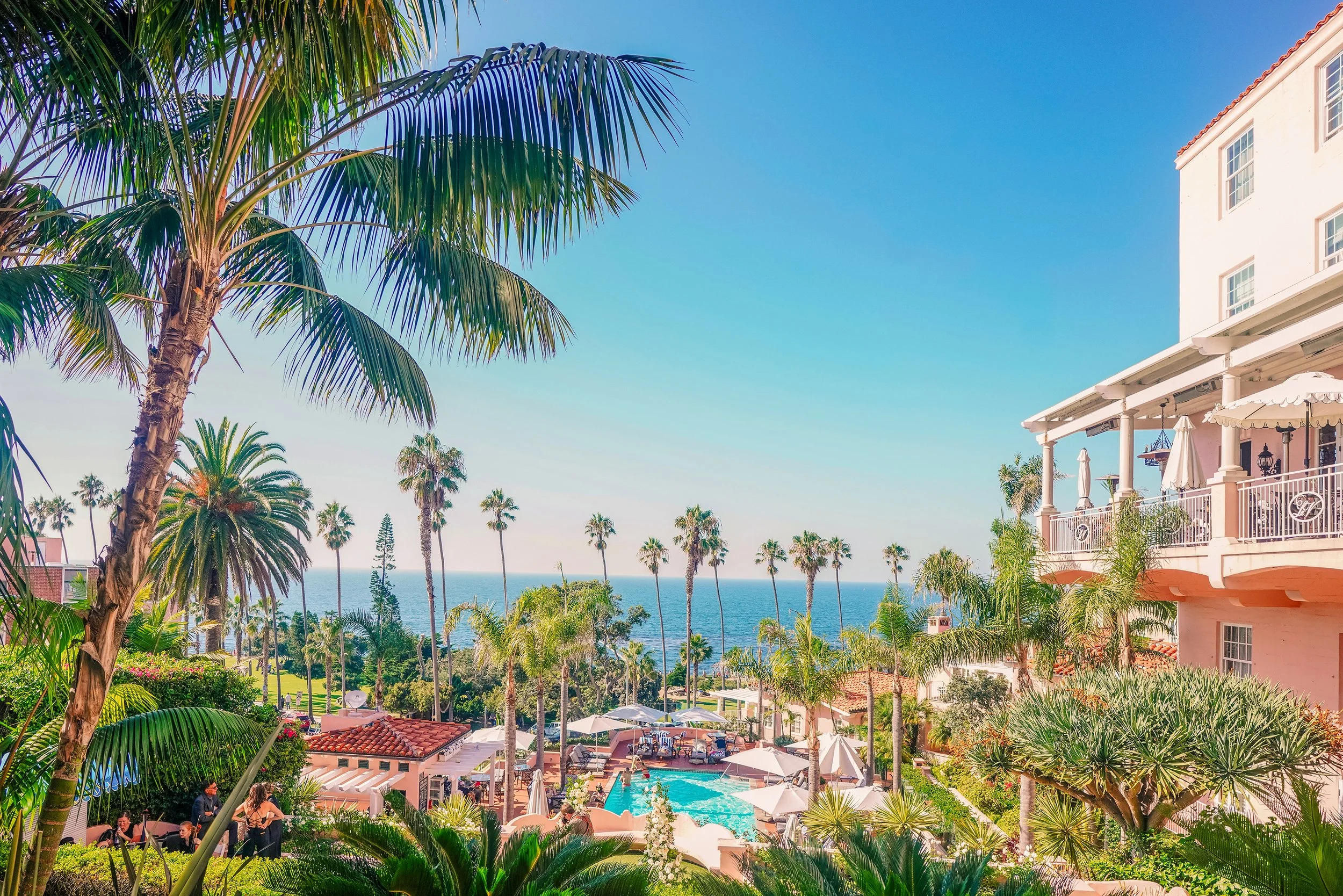 An elevated, daytime view of the multi-story, pink-stucco La Valencia Hotel in La Jolla, California, featuring a swimming pool area surrounded by palm trees and lush greenery, with the Pacific Ocean visible in the background under a blue sky.