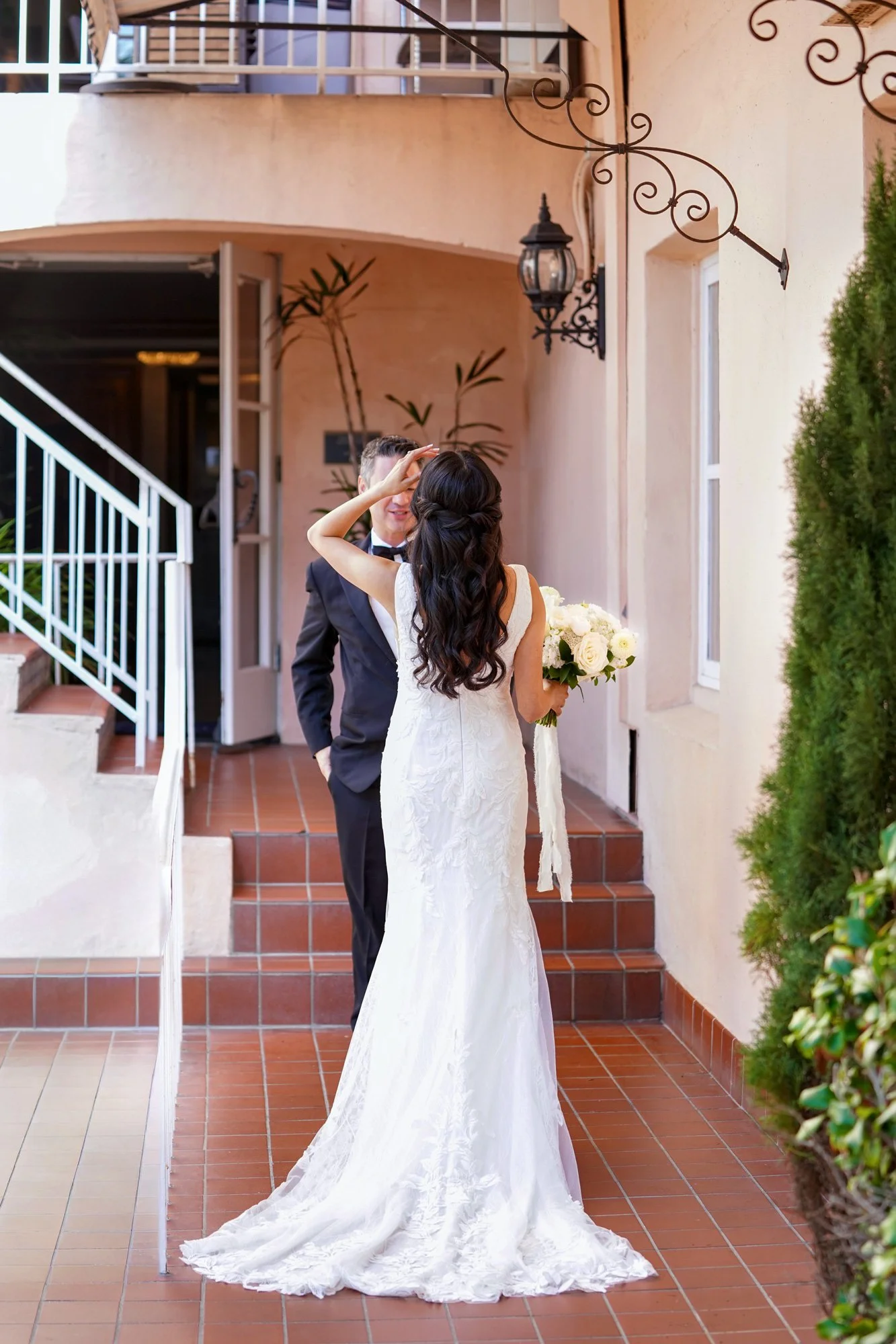 An outdoor photograph capturing a "first look" moment between a bride in a white lace wedding dress holding a white floral bouquet and a groom in a black tuxedo. They are standing on a terracotta-tiled stairway with a white railing, in front of a pin