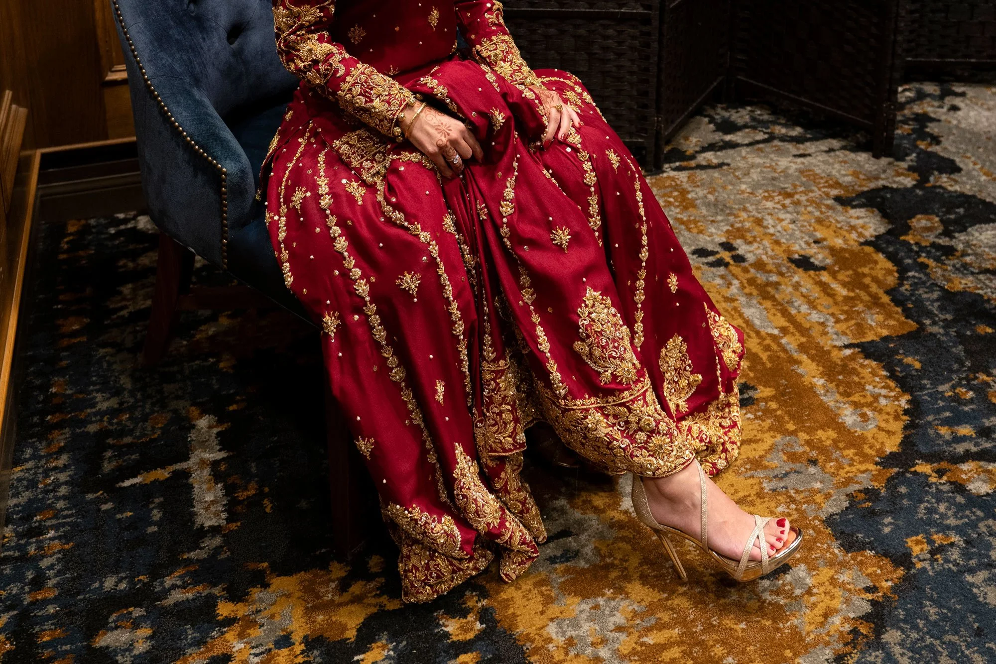 A close-up, indoor photograph of a person seated in a blue velvet chair, wearing an ornate, full-length deep red dress with elaborate gold embroidery. The person is also wearing strappy gold high heels with red pedicure and their hands display light 