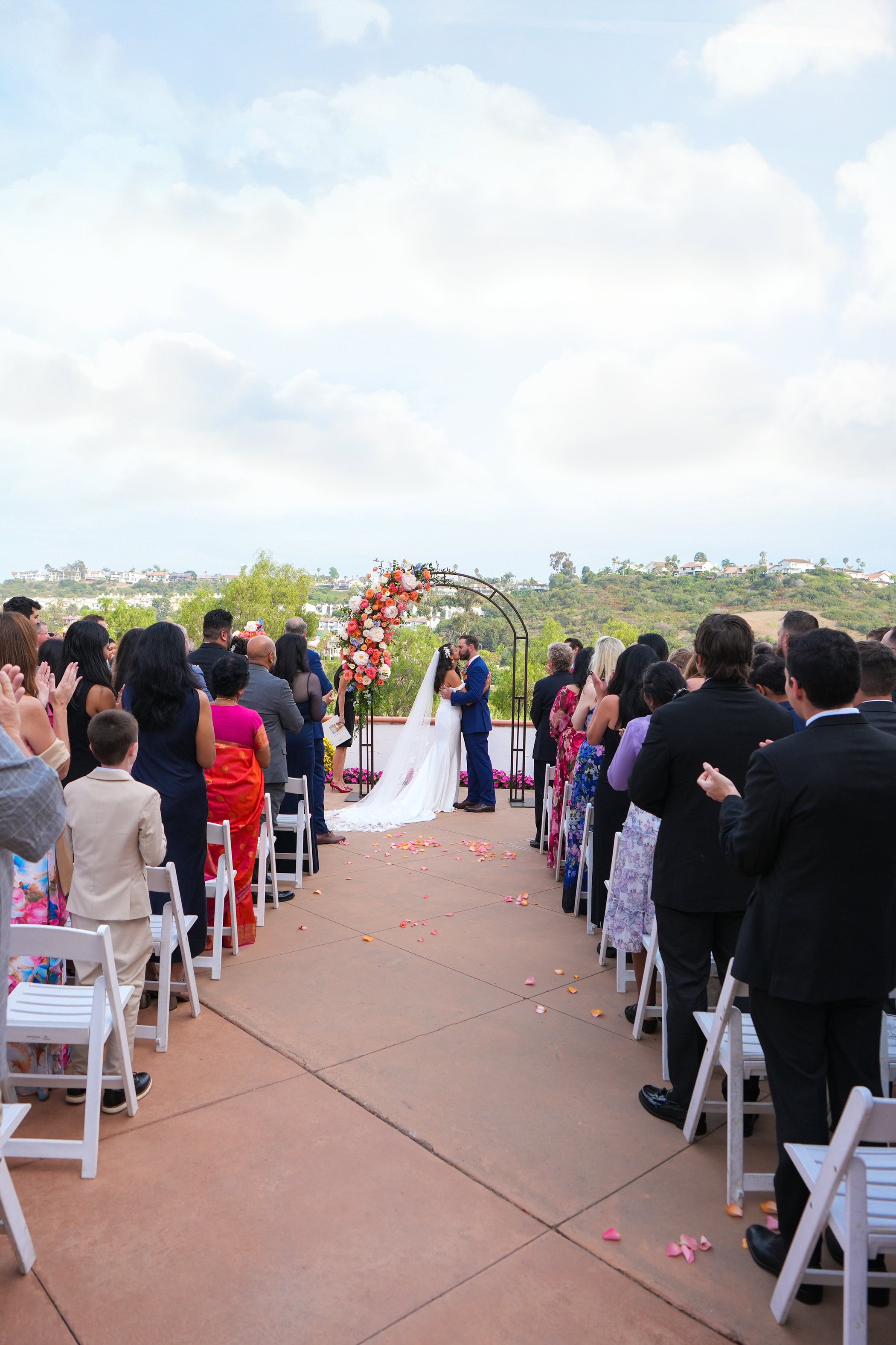 A wide-angle wedding ceremony photograph by Reimo Photography taken from the back of the aisle. A bride and groom are sharing their first kiss under a black metal arch adorned with vibrant orange and pink flowers. Guests seated in white chairs on a t