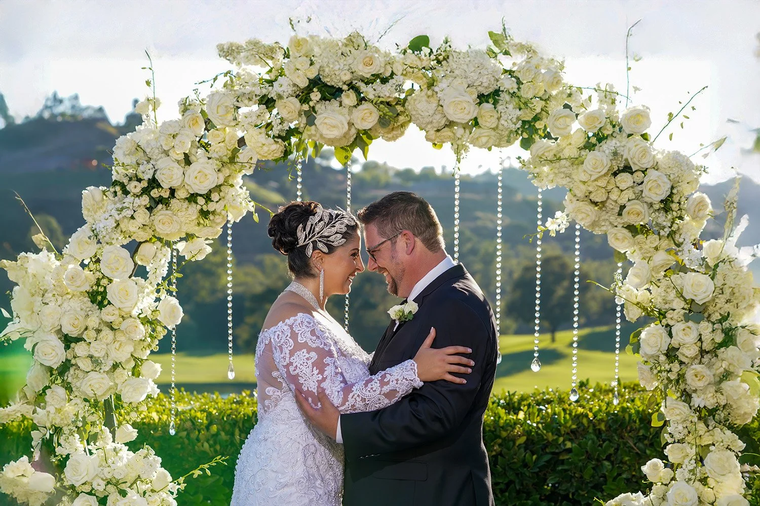 A bride and groom smile at each other, foreheads touching, beneath a lush white floral arch. The bride wears a long-sleeved lace gown and an ornate silver headpiece, while the groom is in a black tuxedo. Crystal strands hang from the arch against a b