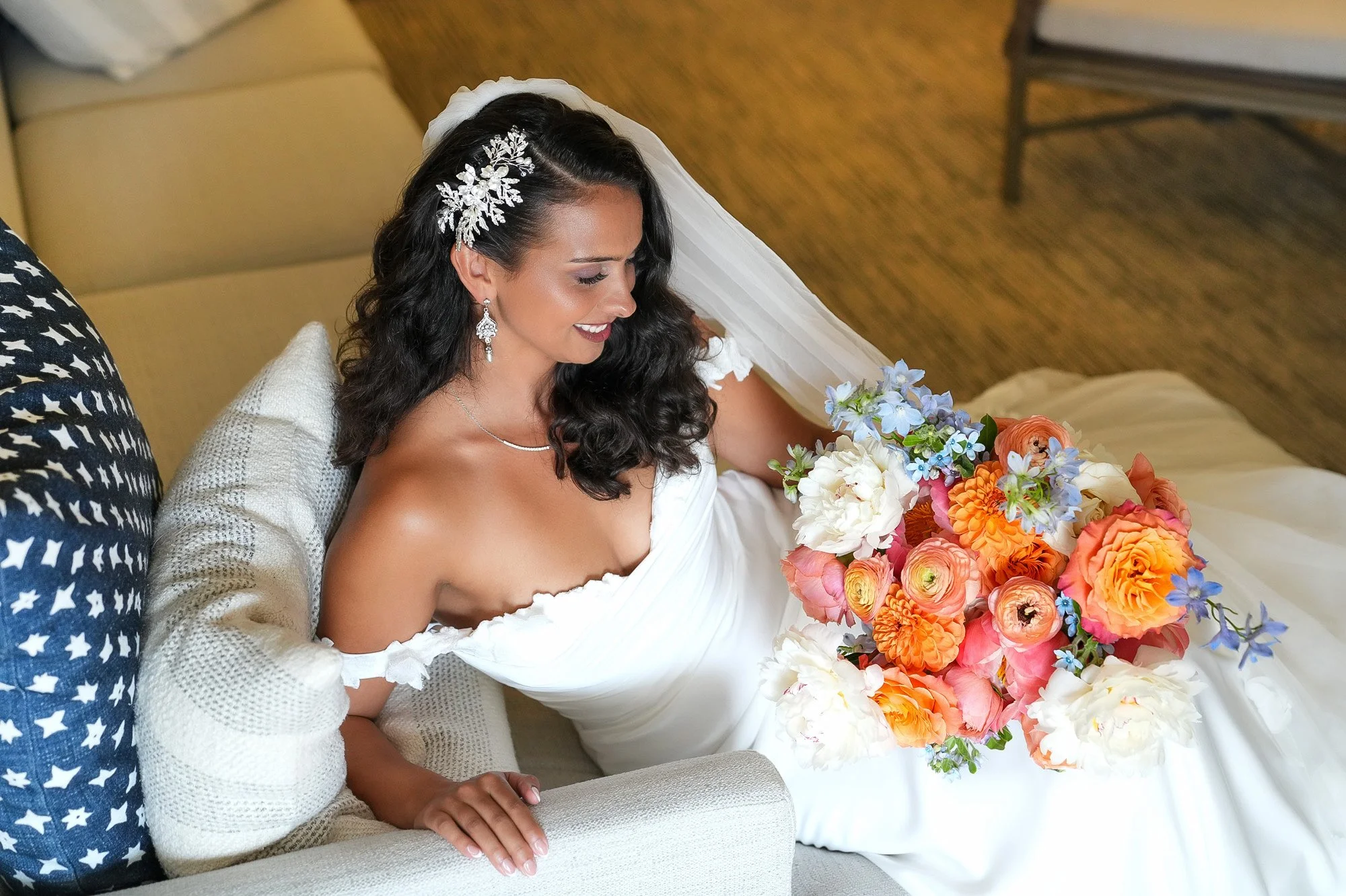 A smiling bride with dark, curly hair wearing a white, off-the-shoulder gown and a floral hairpiece sits on a couch with a large bouquet of orange, pink, blue, and white flowers.


