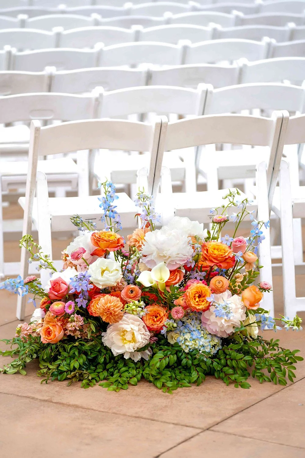 A vibrant floor-level floral arrangement featuring orange, white, pink, and blue flowers set in front of rows of white wedding ceremony chairs on a tiled floor.