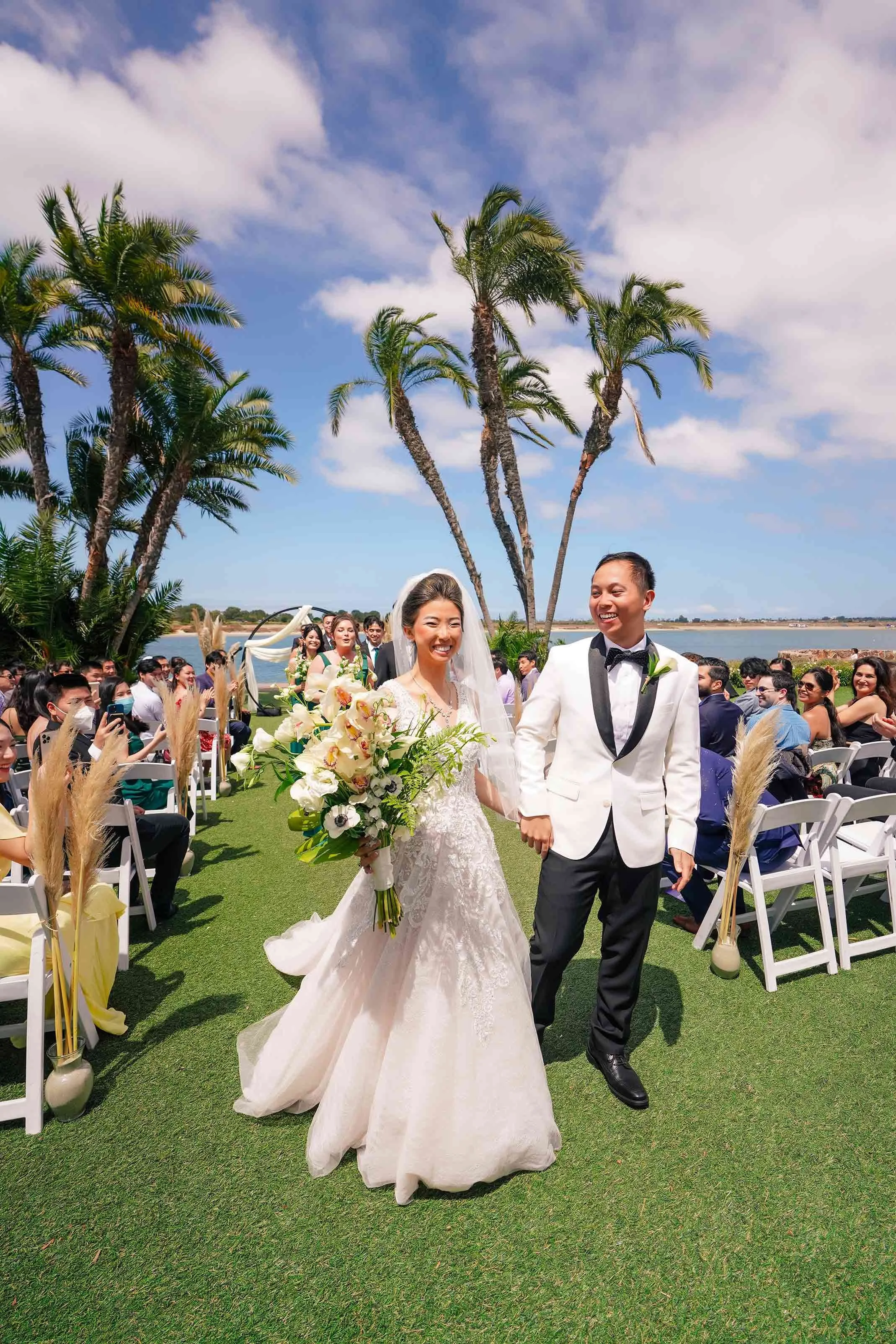A smiling bride in a white lace gown with a large bouquet and a groom in a white tuxedo jacket walk hand-in-hand down an outdoor wedding aisle lined with palm trees and guests in San Diego.