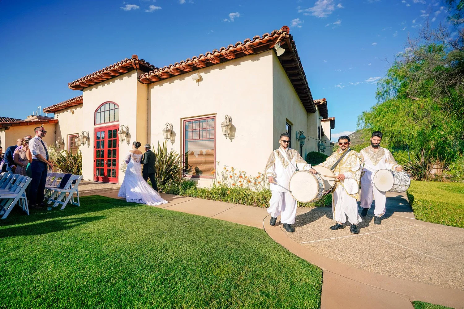 A wedding procession on a sunny day at the Maderas Golf Club in Poway, California. Three dhol players in traditional white and gold attire lead the procession with their drums along a curved walkway. Behind them, a bride in a white wedding dress and 