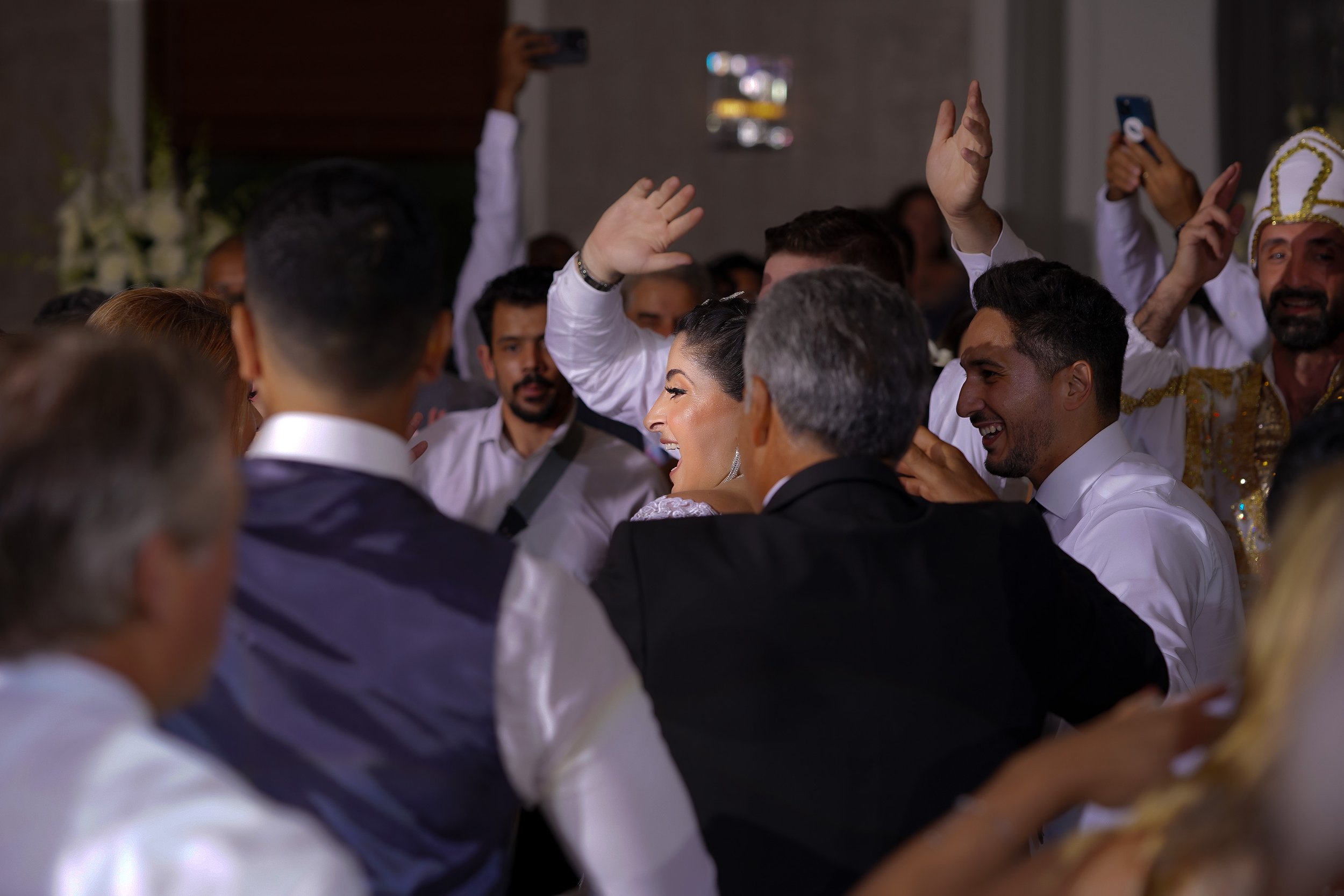 A joyful bride and groom celebrate on a crowded dance floor surrounded by cheering guests. The bride, seen in profile with a wide smile, raises her hands while dancing in her wedding gown. Guests around them are laughing and clapping, with some holdi