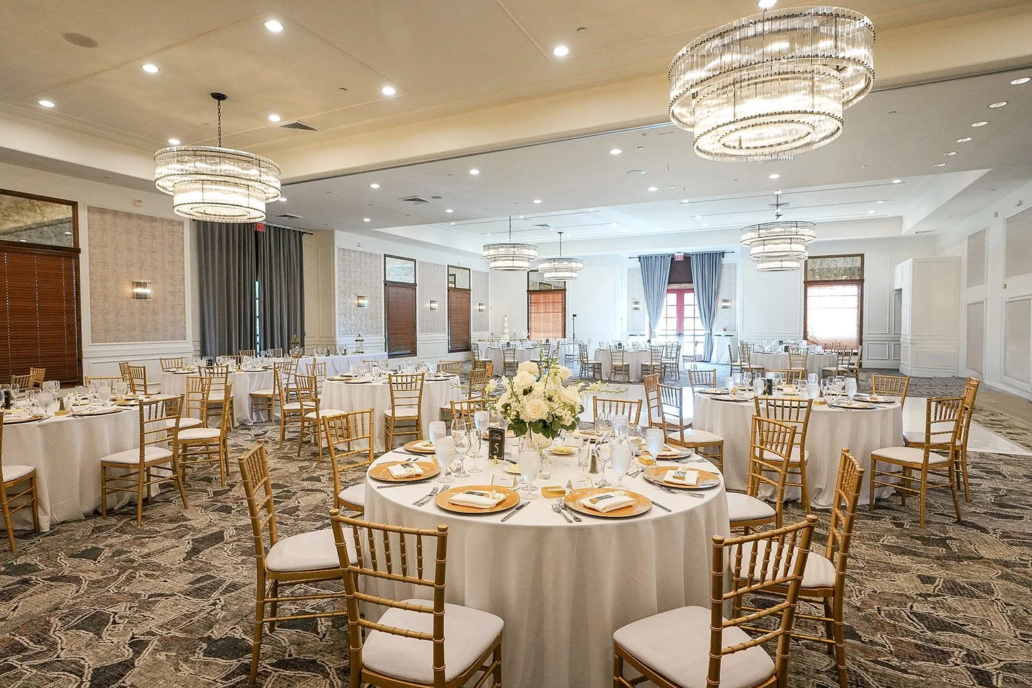 An elegant wedding reception setup in a banquet hall. Round tables are set with white linens, gold charger plates, and white floral centerpieces. Gold Chiavari chairs surround the tables, and large, modern crystal chandeliers hang from the ceiling. T