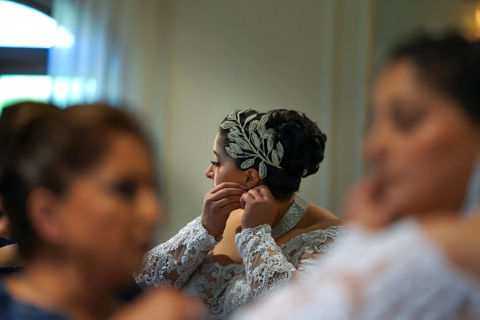 A bride in an intricate lace dress puts on her earrings while getting ready for her wedding. She has her hair in a dark updo adorned with a large, sparkling silver leaf-shaped headpiece. The foreground is softly blurred with the figures of other peop
