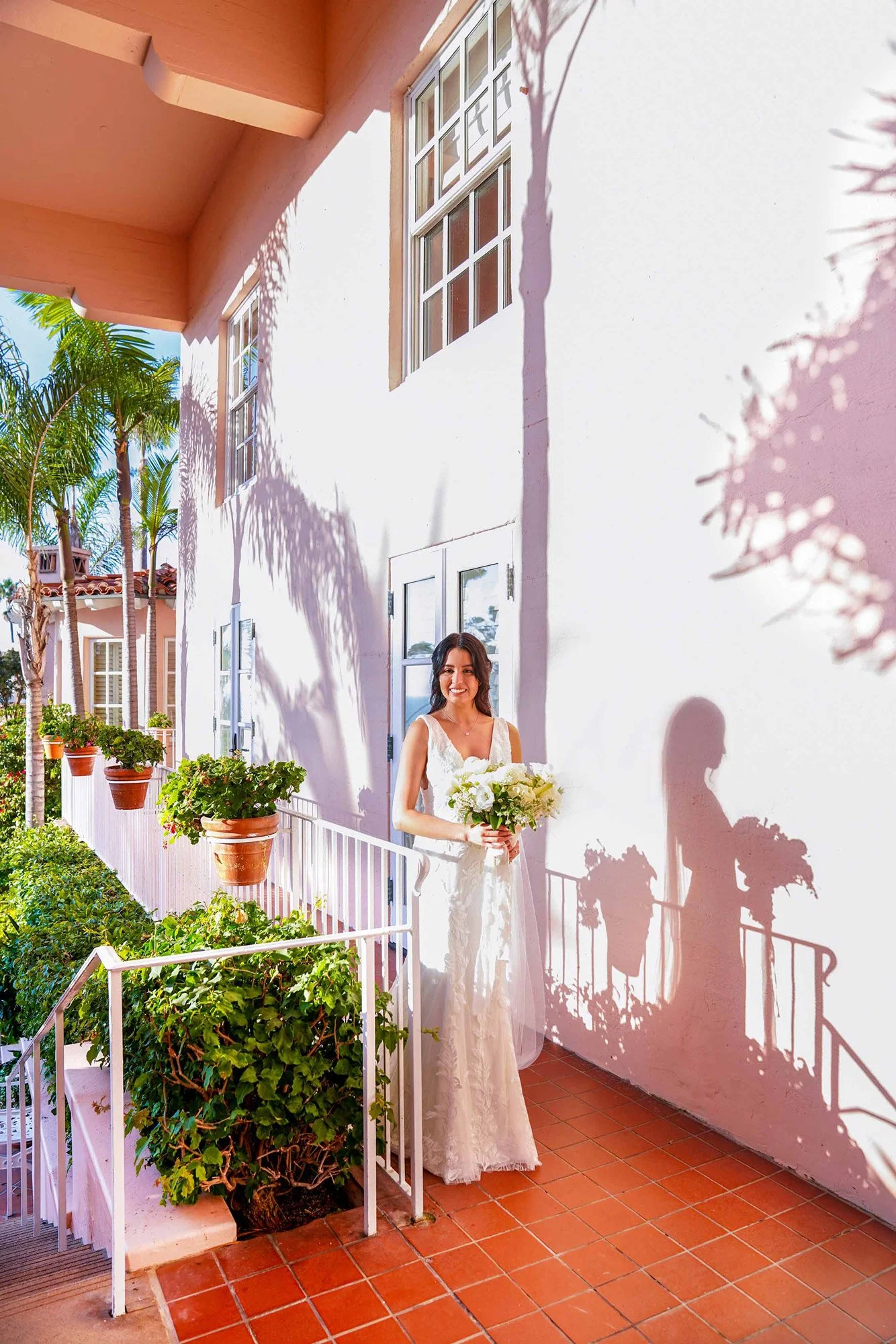 A full-length outdoor photograph of a smiling bride in a white lace wedding dress, holding a large white floral bouquet. She is standing on a red-tiled patio of a pink stucco building, with strong sunlight casting prominent shadows of her, the railin
