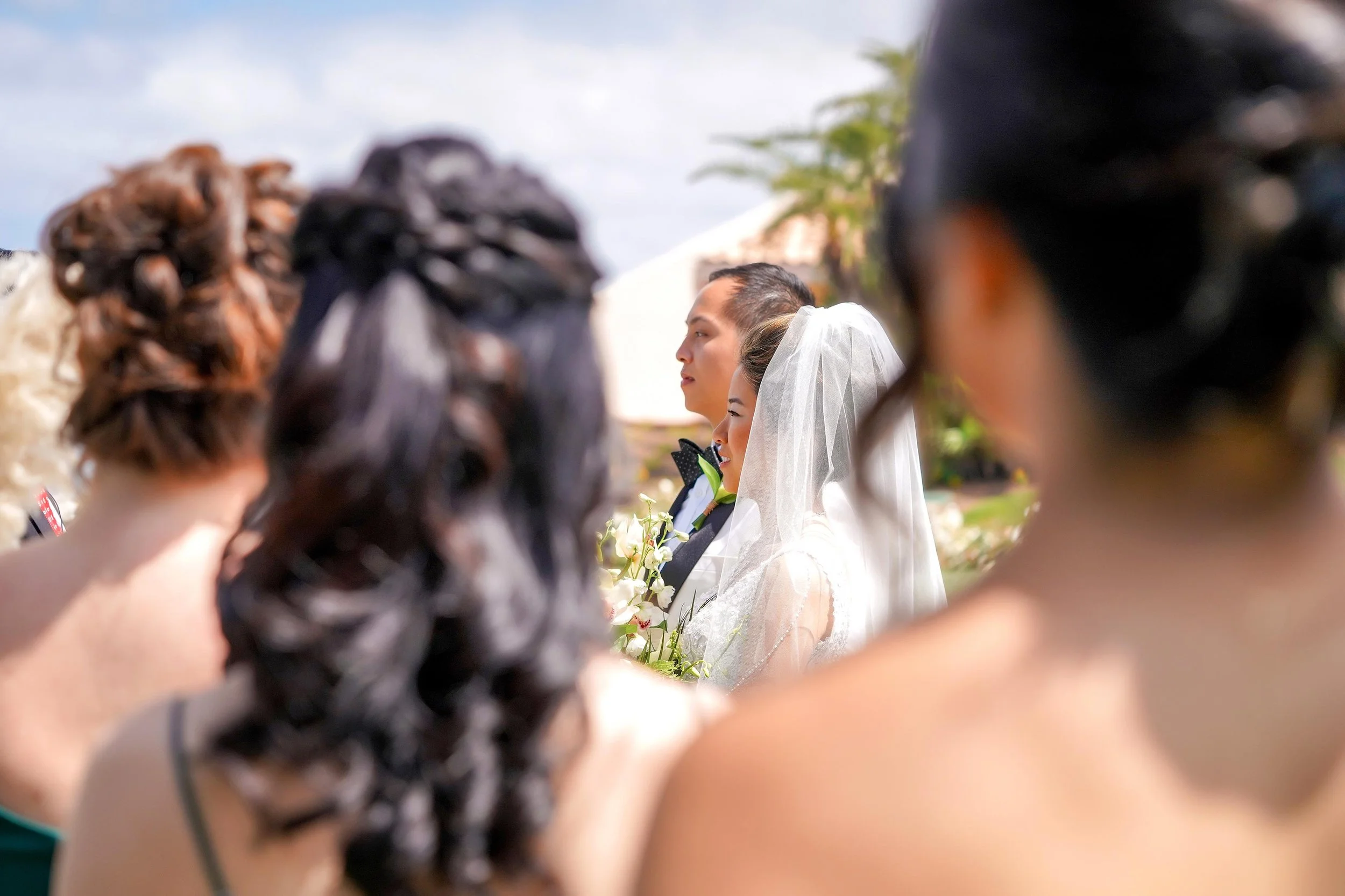 A candid, shallow depth-of-field photograph taken from the perspective of the wedding party, showing the profiles of a bride in a white veil and a groom in a black tuxedo jacket standing at the altar during a sunny outdoor ceremony.


