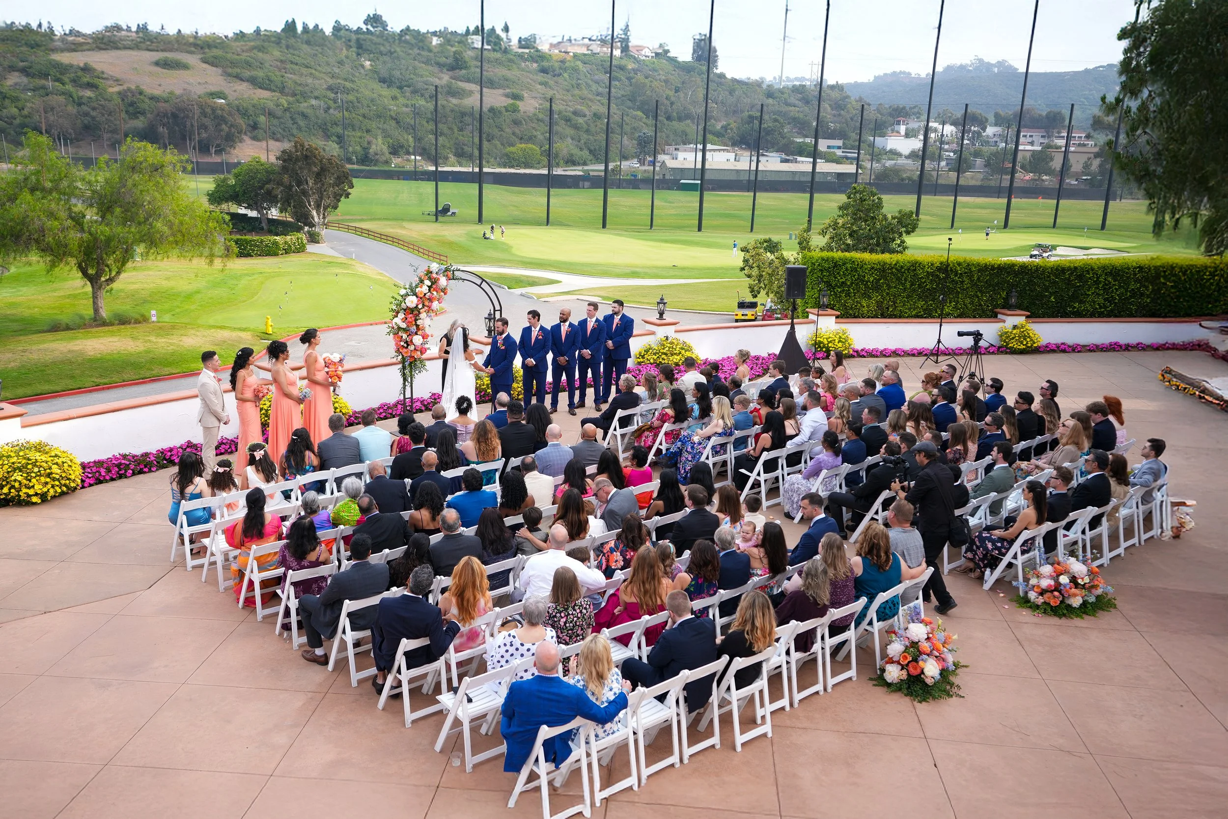 A wide-angle, high-angle outdoor wedding ceremony photo by Reimo Photography at Omni resort, LA Costa. Guests are seated in white folding chairs on a paved terrace, facing a couple at a floral-adorned arch. The ceremony overlooks the rolling green fa