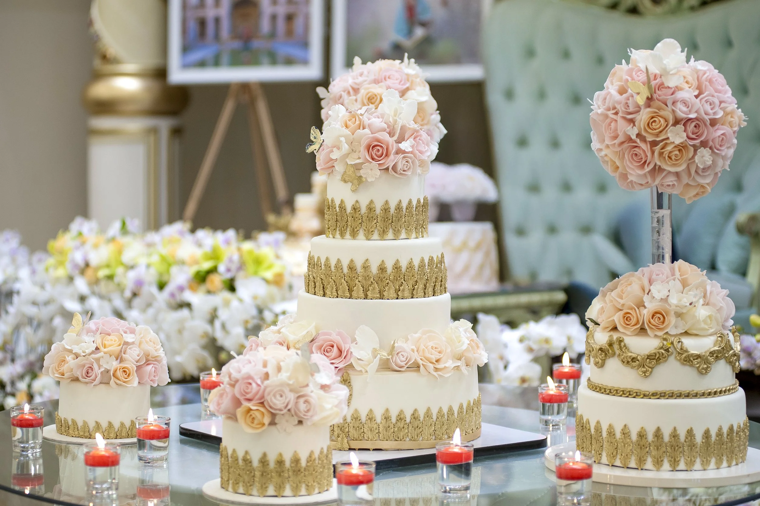 A five-tiered white and gold wedding cake and several smaller matching single-tiered cakes, all decorated with pink and white roses, displayed on a glass table surrounded by red votive candles.
