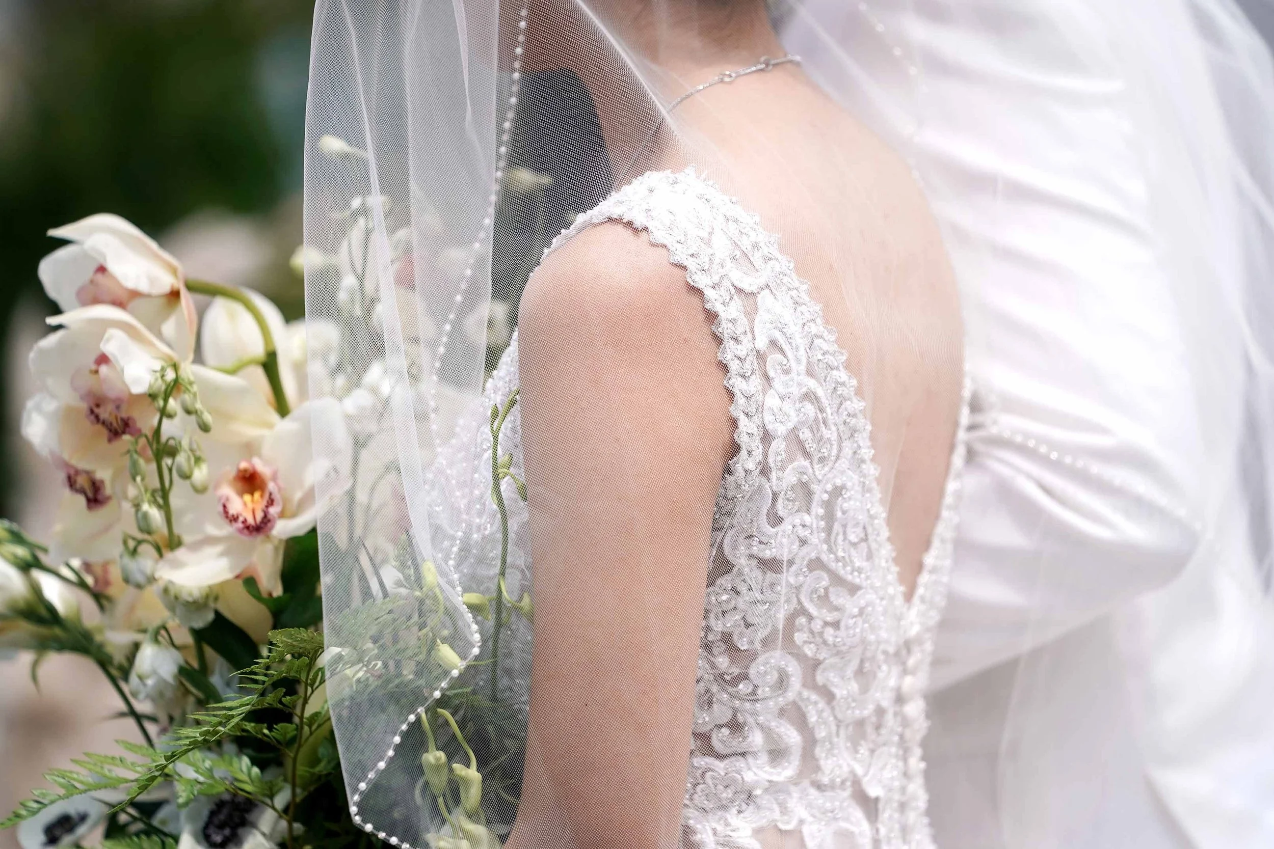 A close-up photograph of a bride from behind, showcasing the intricate white lace pattern on her wedding dress, a sheer veil with beaded trim, and a bouquet of white orchids in the foreground.