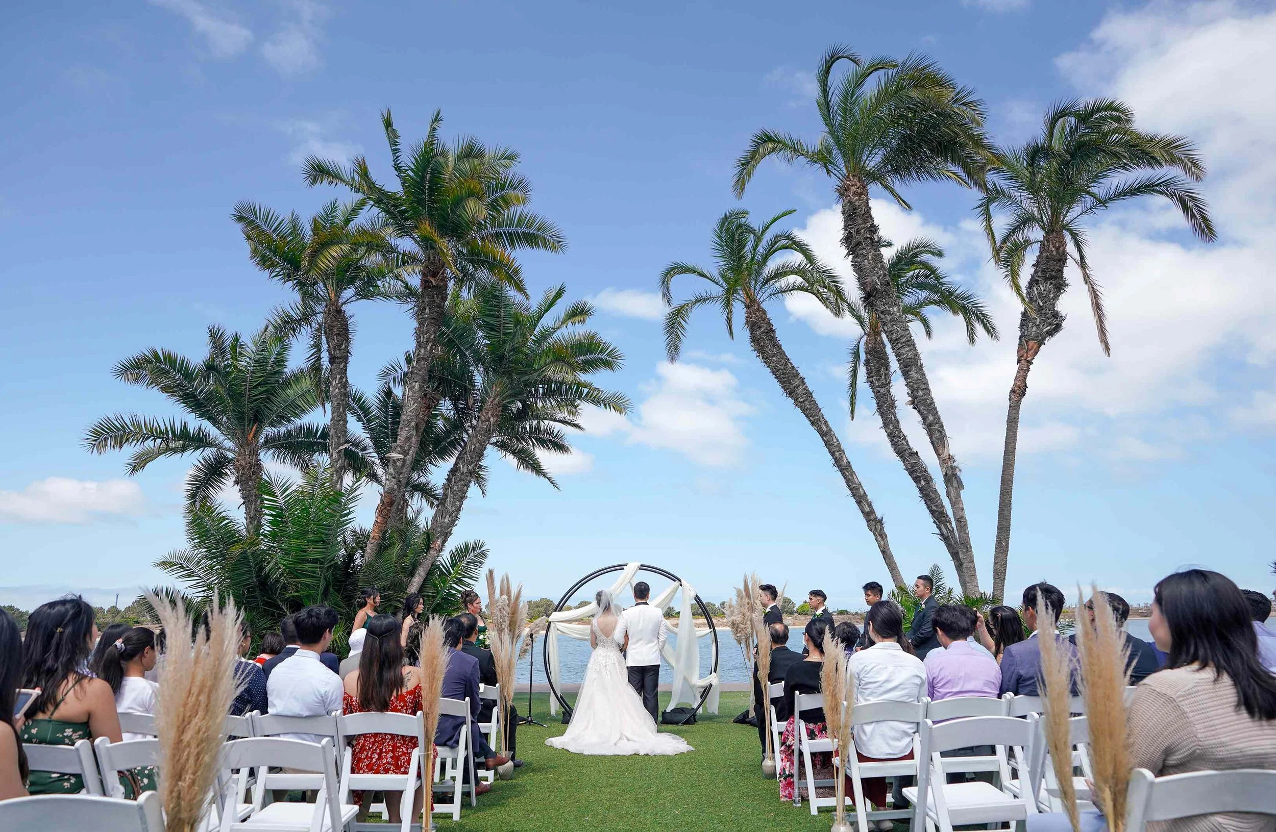 A sunny outdoor wedding ceremony overlooking the water in Mission Bay, San Diego, featuring the bride and groom at the altar under a circular arch, framed by tall palm trees.