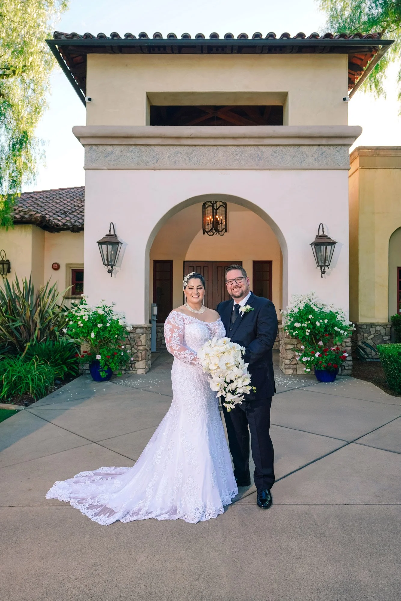 A full-length outdoor photograph of a smiling bride in a white lace wedding dress holding a large white bouquet, posing with the groom in a black suit in front of a Spanish-style building entrance of Maderas Golf Club with an archway and lanterns.


