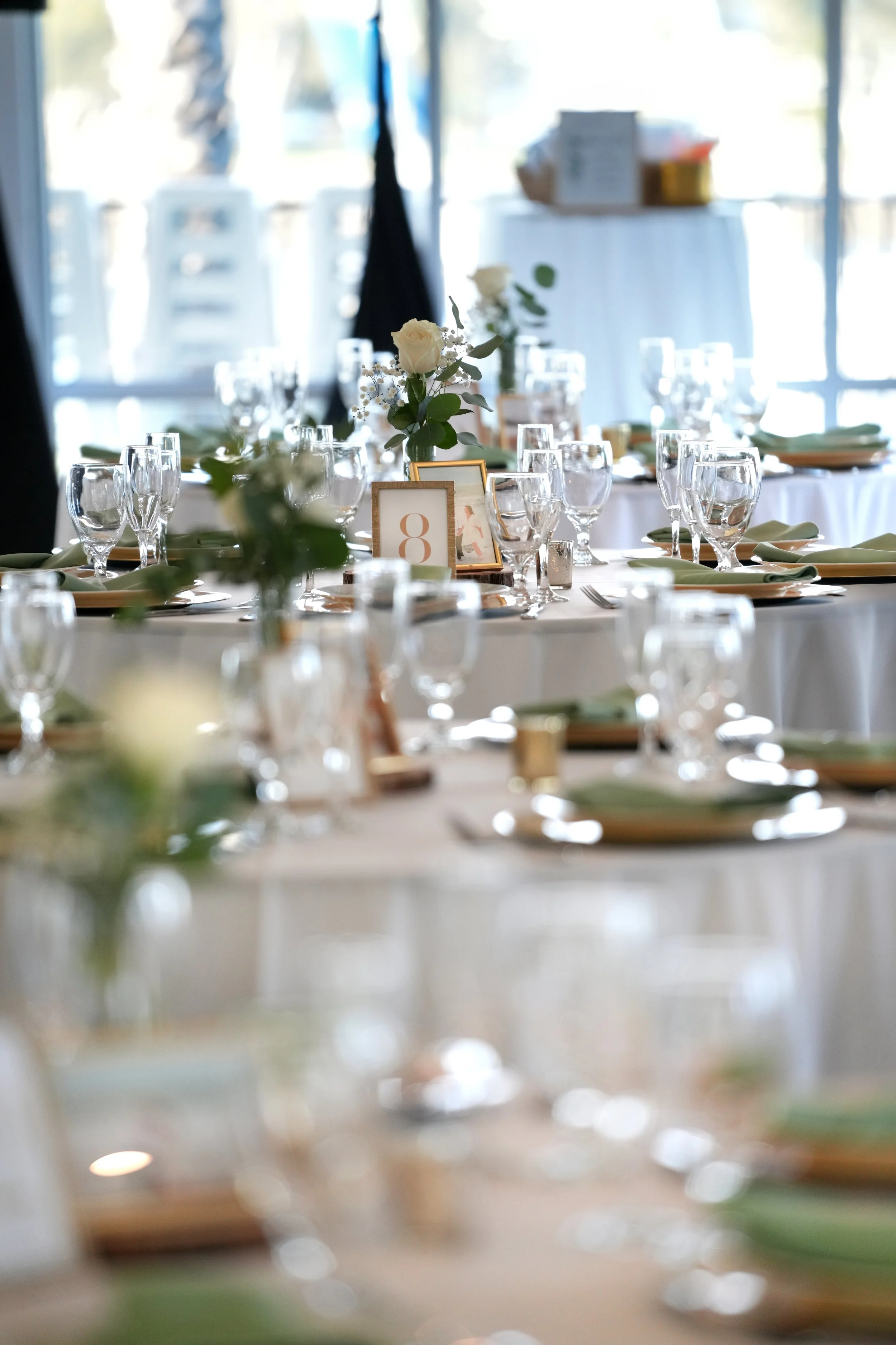 A formal dining table set for an event with gold charger plates, green cloth napkins, clear glassware, small white rose centerpieces, and a gold-framed table number "8".