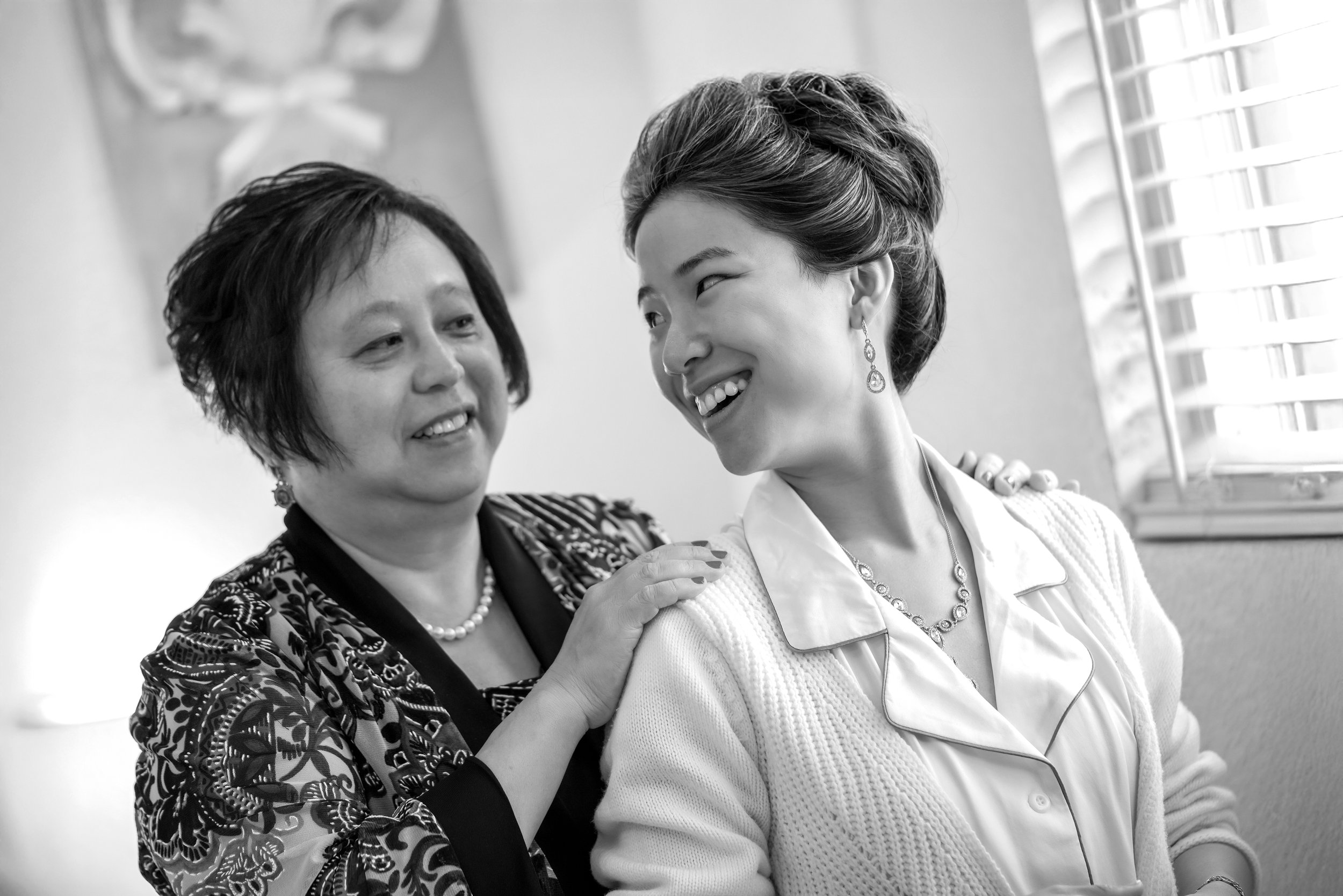 Mother and daughter smiling warmly at each other. The woman on the left has short dark hair and is wearing a patterned top and a pearl necklace. She has her hand on the shoulder of the younger woman on the right. The younger woman has her hair styled