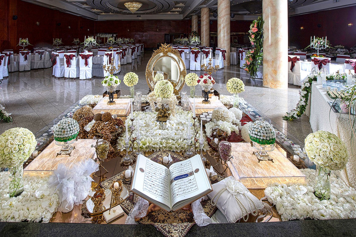 An elaborate, traditional Persian wedding table setting (Sofreh Aghd) featuring an ornate gold mirror, candelabras, a holy book, nuts, sweets, and flowers on a white lace tablecloth.