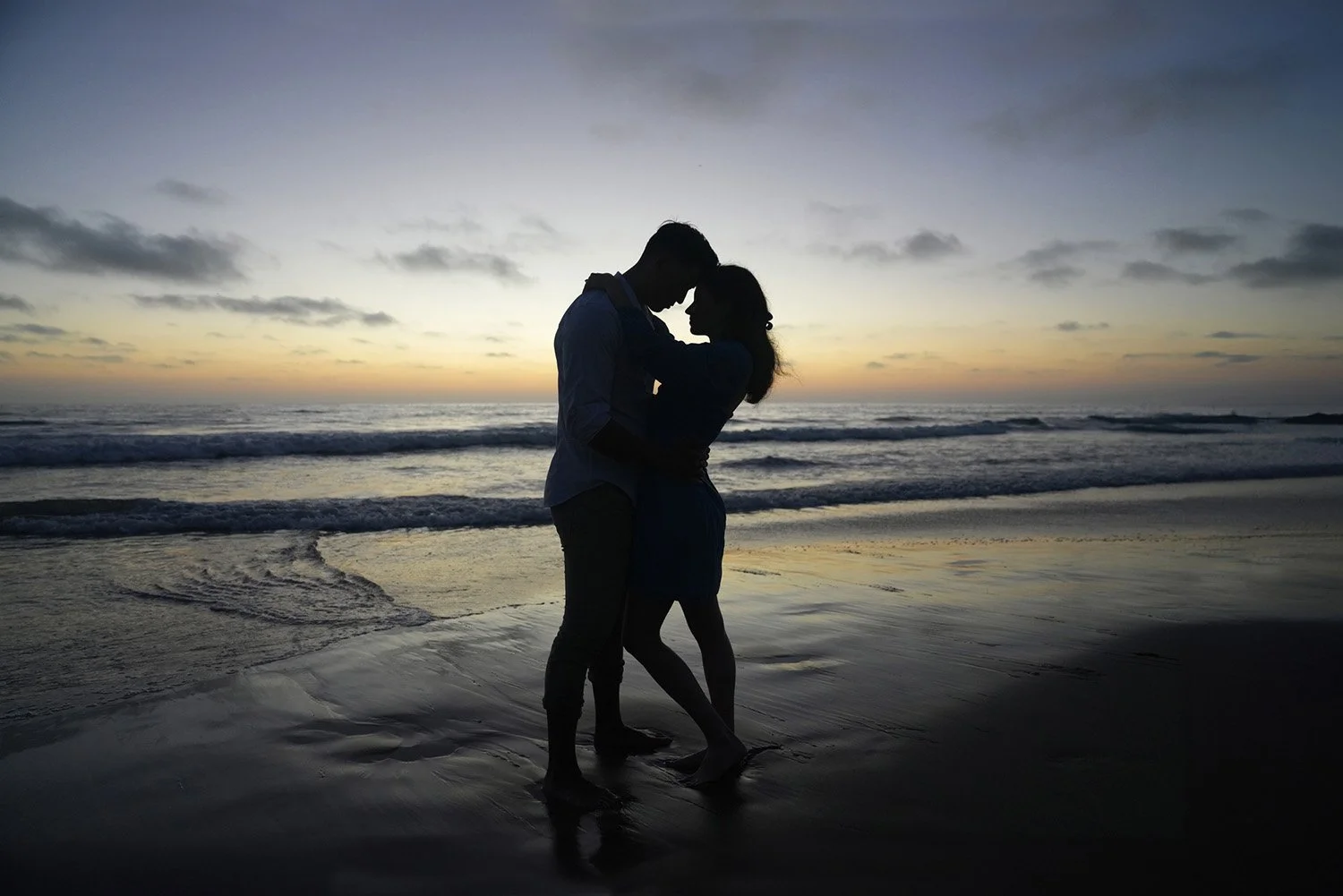 A silhouette photograph of a couple embracing and kissing on a wet sandy beach at sunset or sunrise. The man wears a long-sleeved shirt and pants, and the woman wears a dress. They stand with their arms around each other, silhouetted against the brig