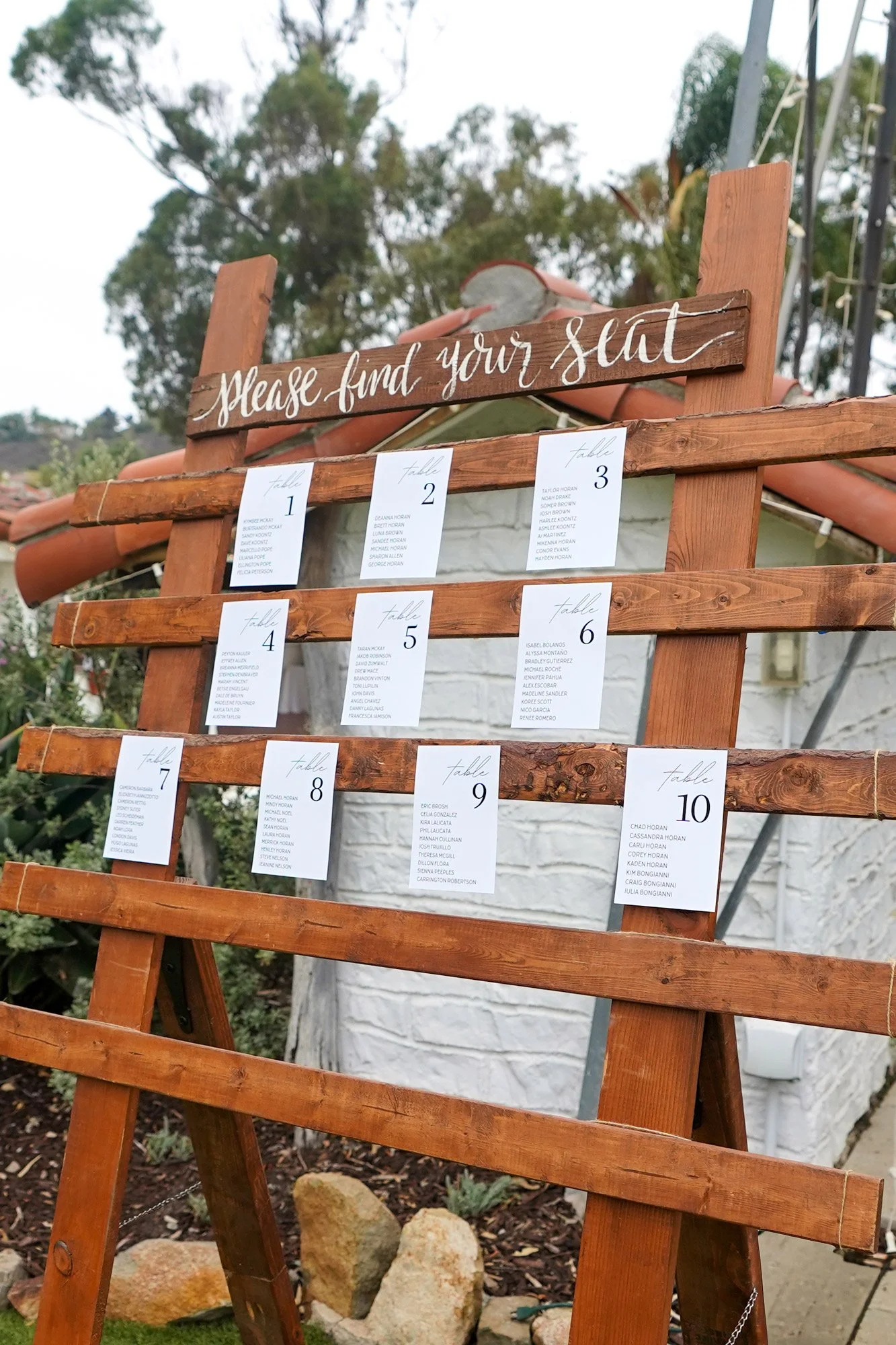 A photo of a rustic wooden seating chart stand outdoors, displaying white cards for tables numbered 1 through 10 with guest names, and a sign on top that reads 'Please find your seat'.