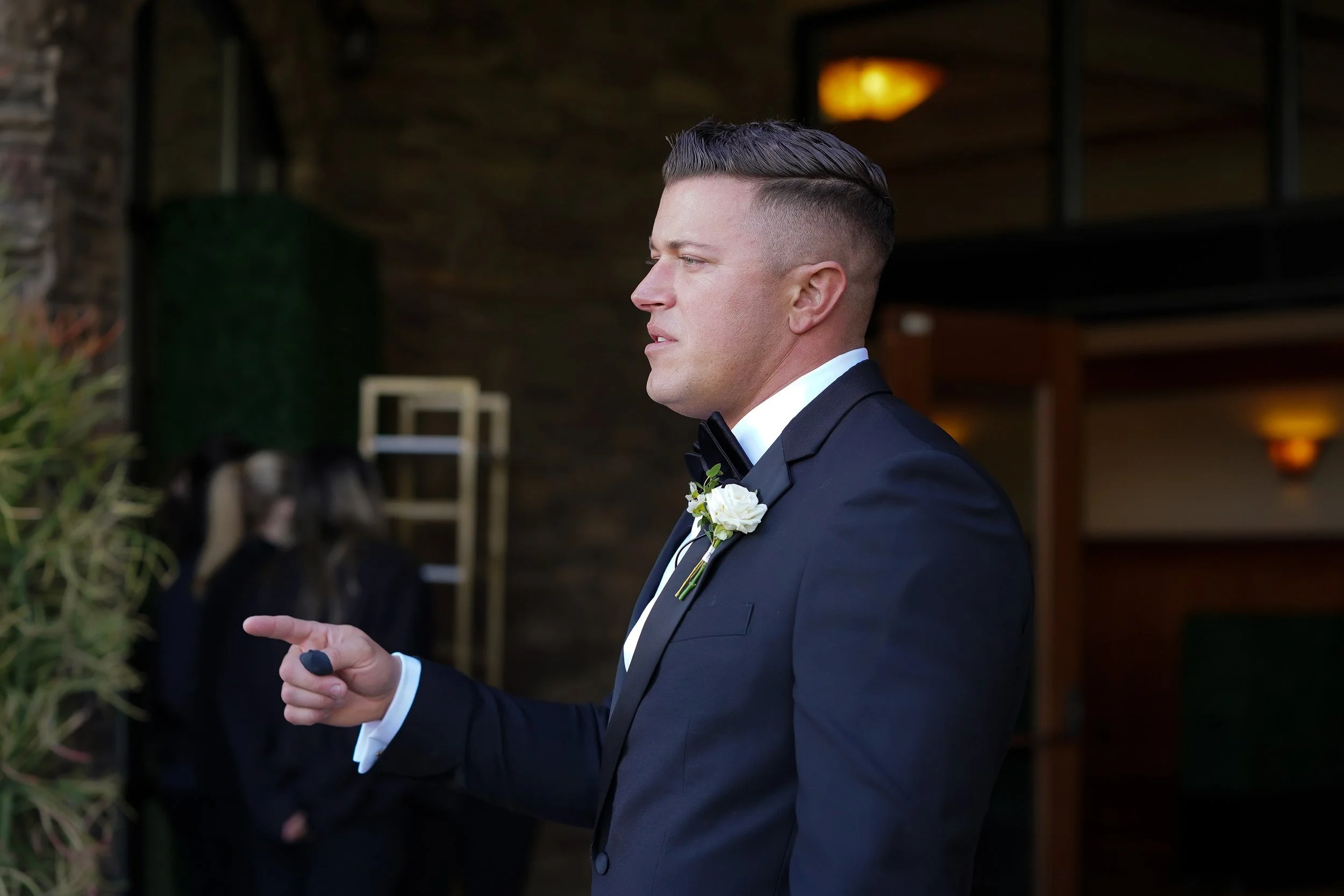 A groom in formal wedding attire pointing his finger while standing near the entrance of an indoor-outdoor reception space.